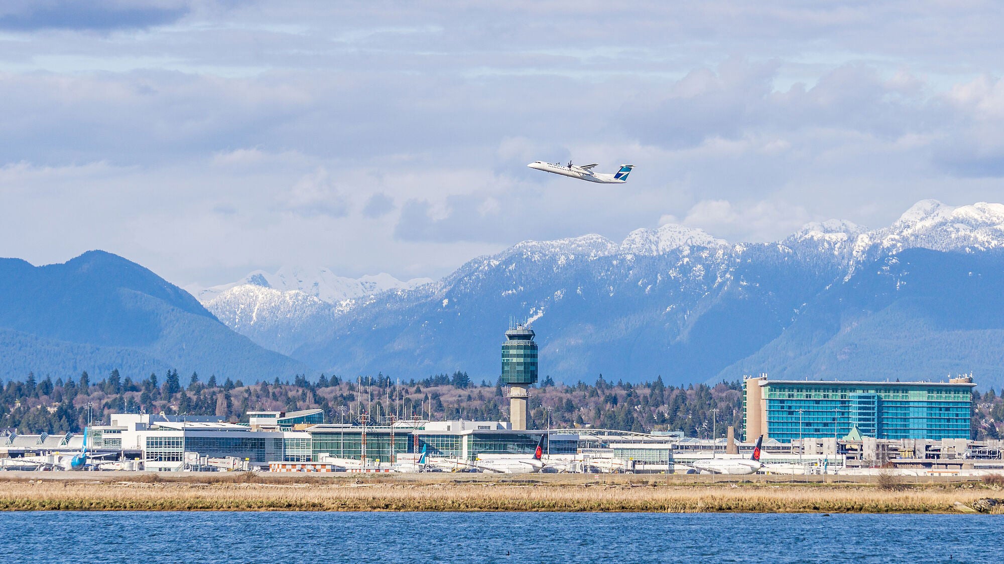 Mountainous backdrop with an airplane taking off over a modern airport. Snow-capped peaks and a cloudy sky convey a serene, expansive atmosphere.