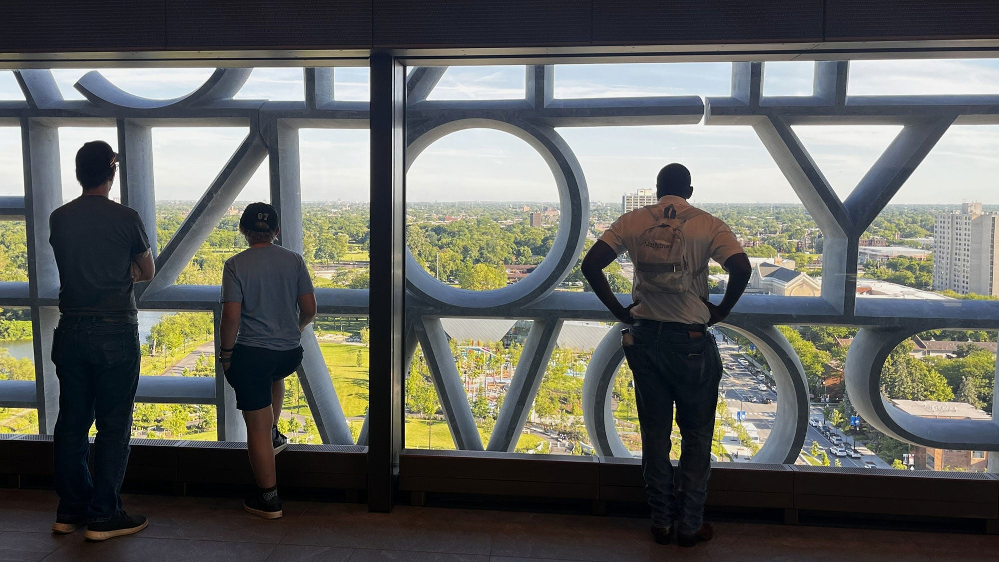 Three visitors looking out from an indoor observation deck at the Obama Presidential Center through large windows with a sculpted concrete letter facade.