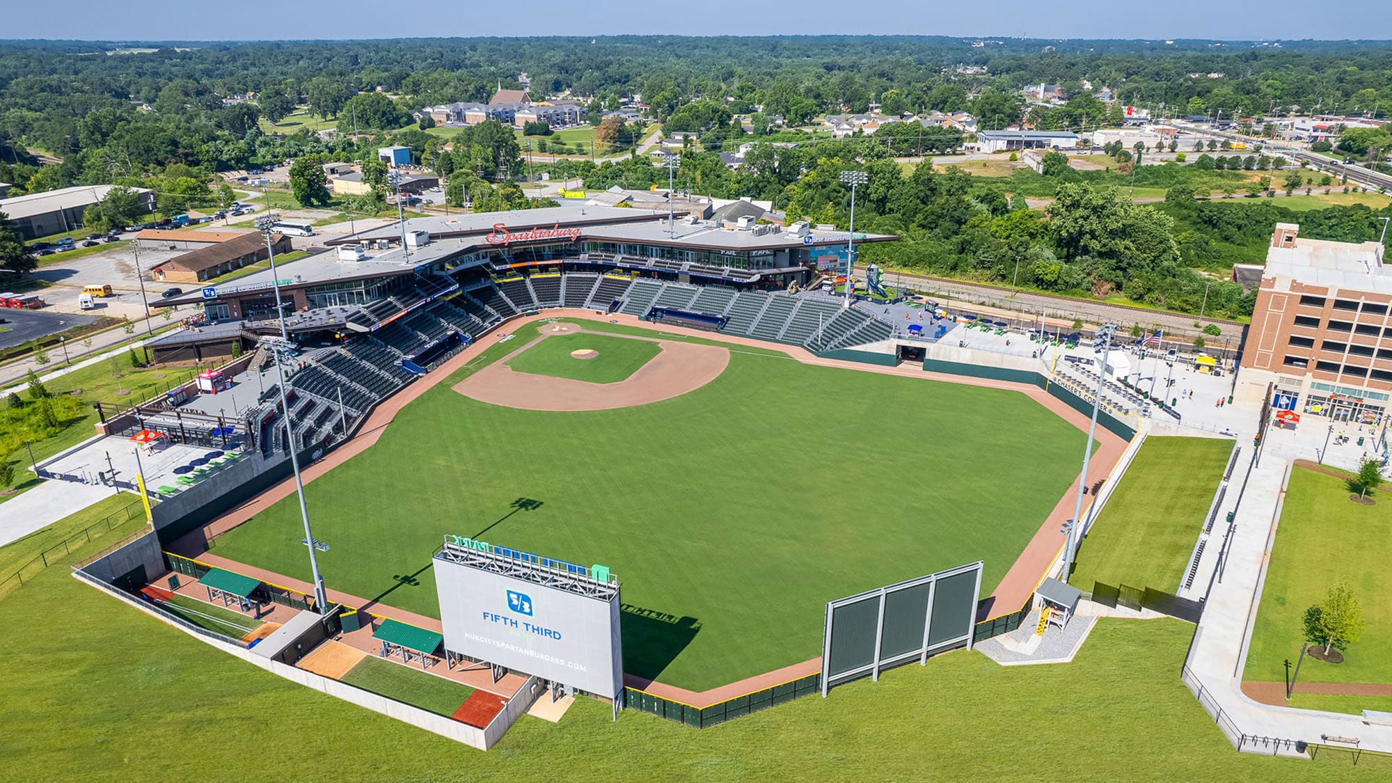A baseball stadium viewed from above, surrounded by green trees and landscaping. 