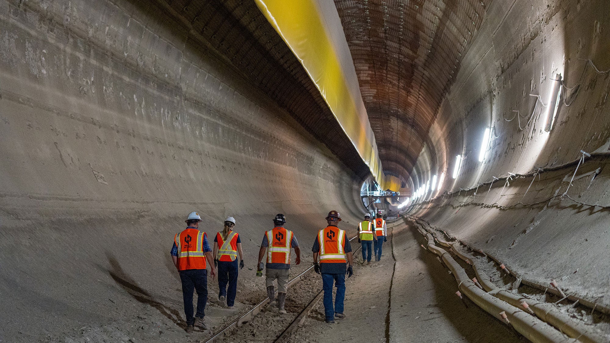 Construction crew wearing high-visibility safety vests walking along tracks inside the Mill Creek underground drainage relief tunnel in Dallas.