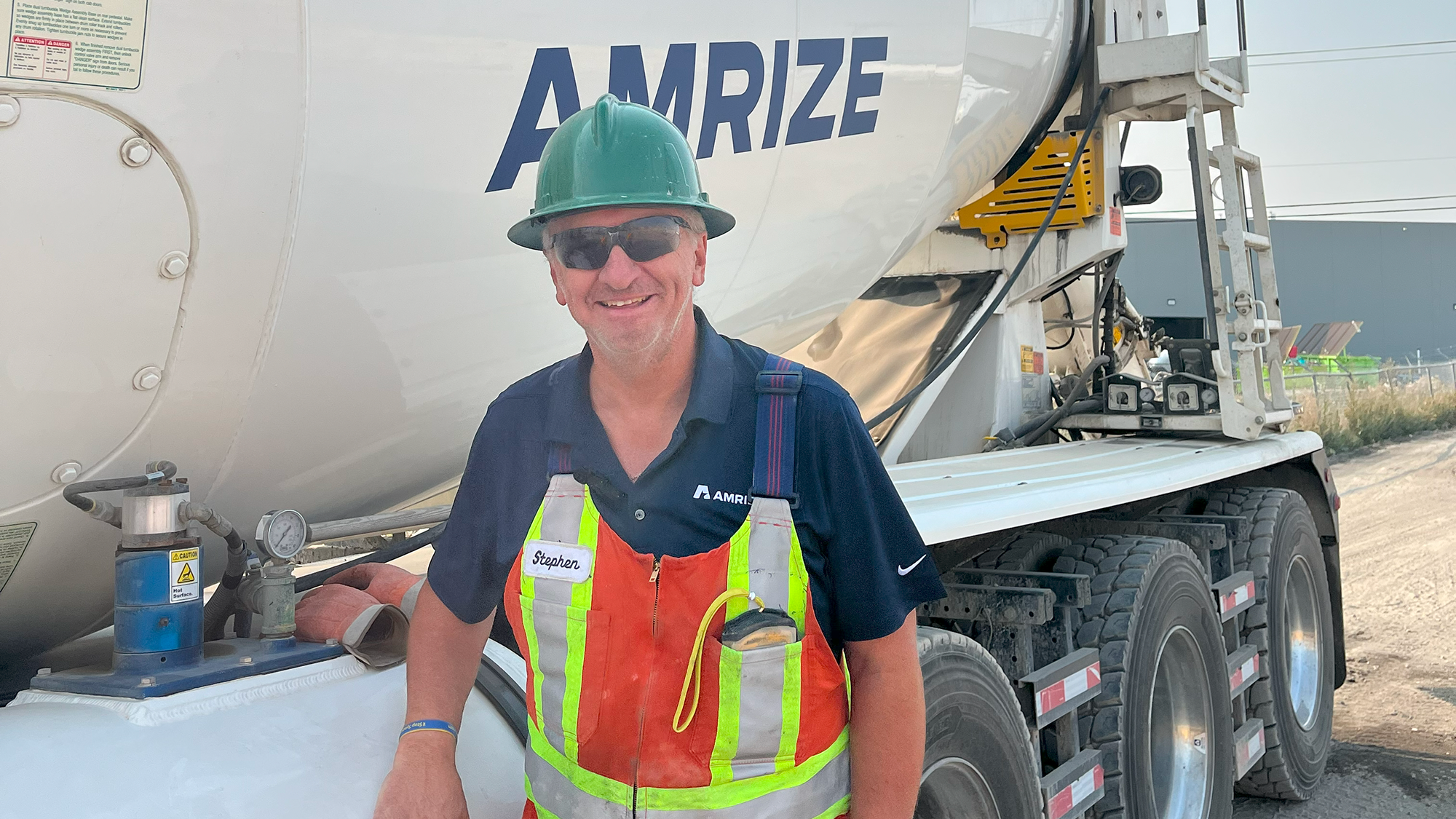 A truck driver in a hard hat and sunglasses stands smiling in front of his truck