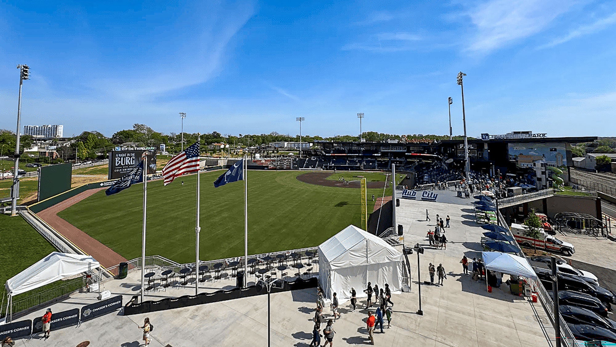 Elevated view of Fifth Third Park minor league baseball stadium in South Carolina, featuring the green outfield. Fans are walking along the concourse under a blue sky. 