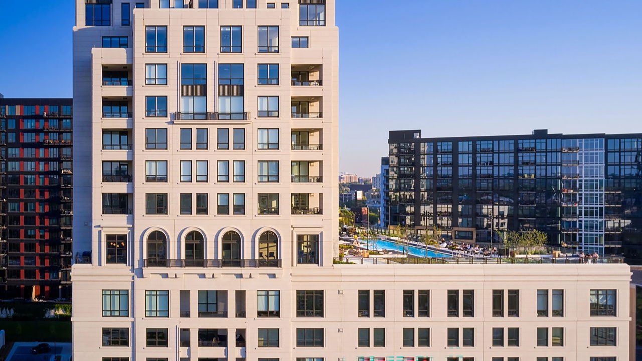 A modern high-rise building, ELEVEN, with large windows and arched features, is set against a clear blue sky. The building’s swimming pool and other nearby condos are visible.
