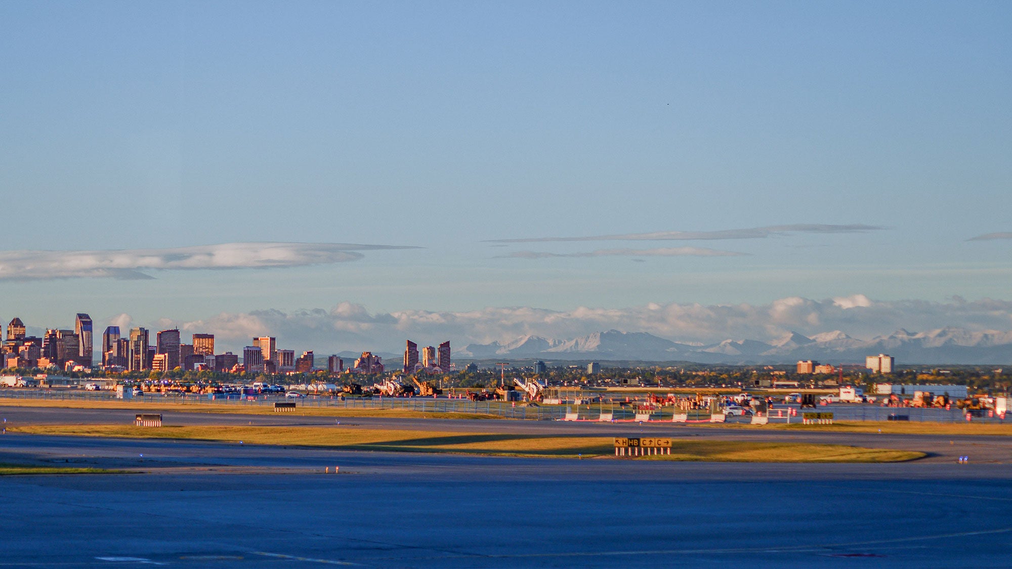 A wide-angle landscape view of the Calgary International Airport tarmac in the foreground, with the Calgary city skyline and the snow-capped Rocky Mountains visible under a clear blue sky in the distance.