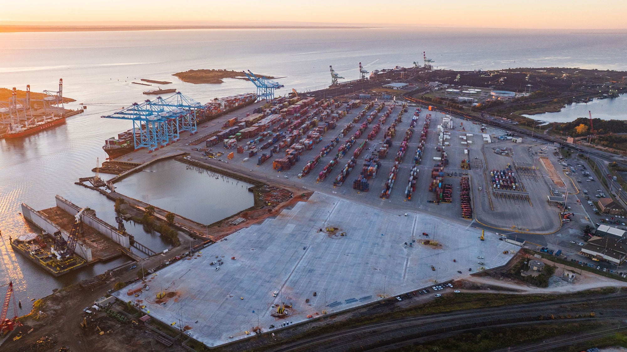 Aerial view of the Port of Mobile at sunset, with numerous shipping containers, blue cranes and calm water.