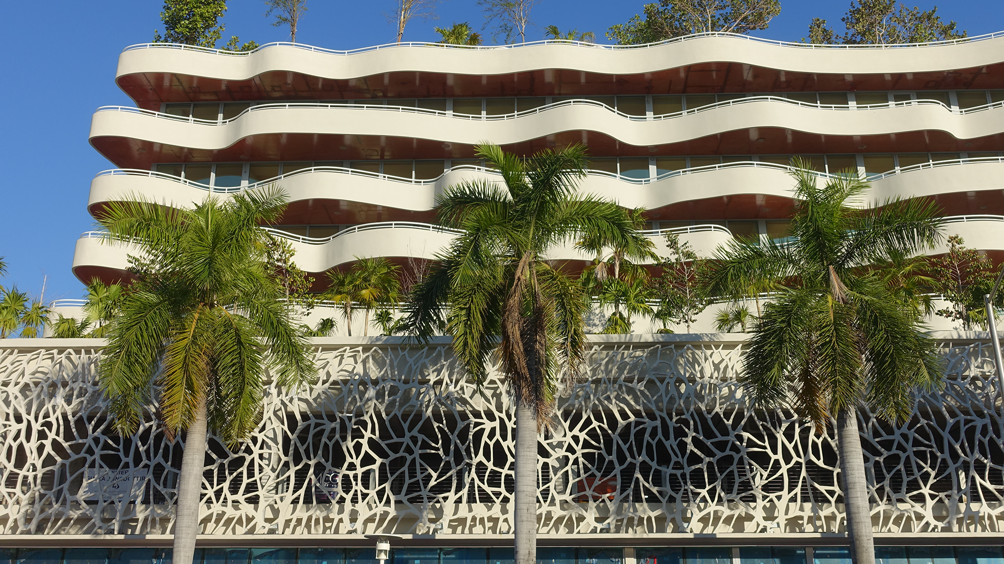 A Miami hotel with a white, wavy facade and balconies is seen from a low angle, with palm trees and a white concrete geometric screen in the foreground. Photo courtesy of Hubert Chang, TYLin.