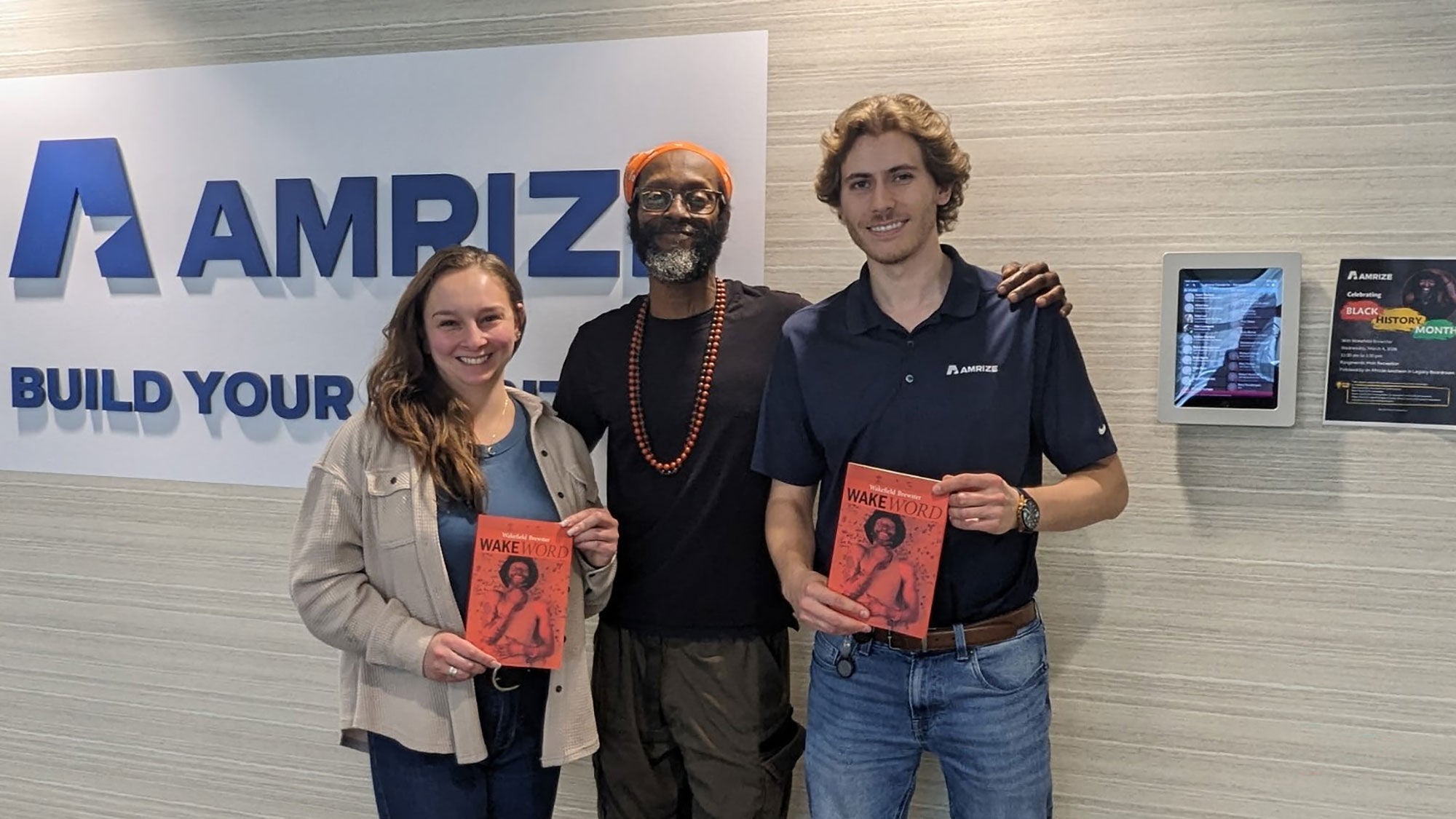 Three people stand together in front of an Amrize company sign holding books of poetry.