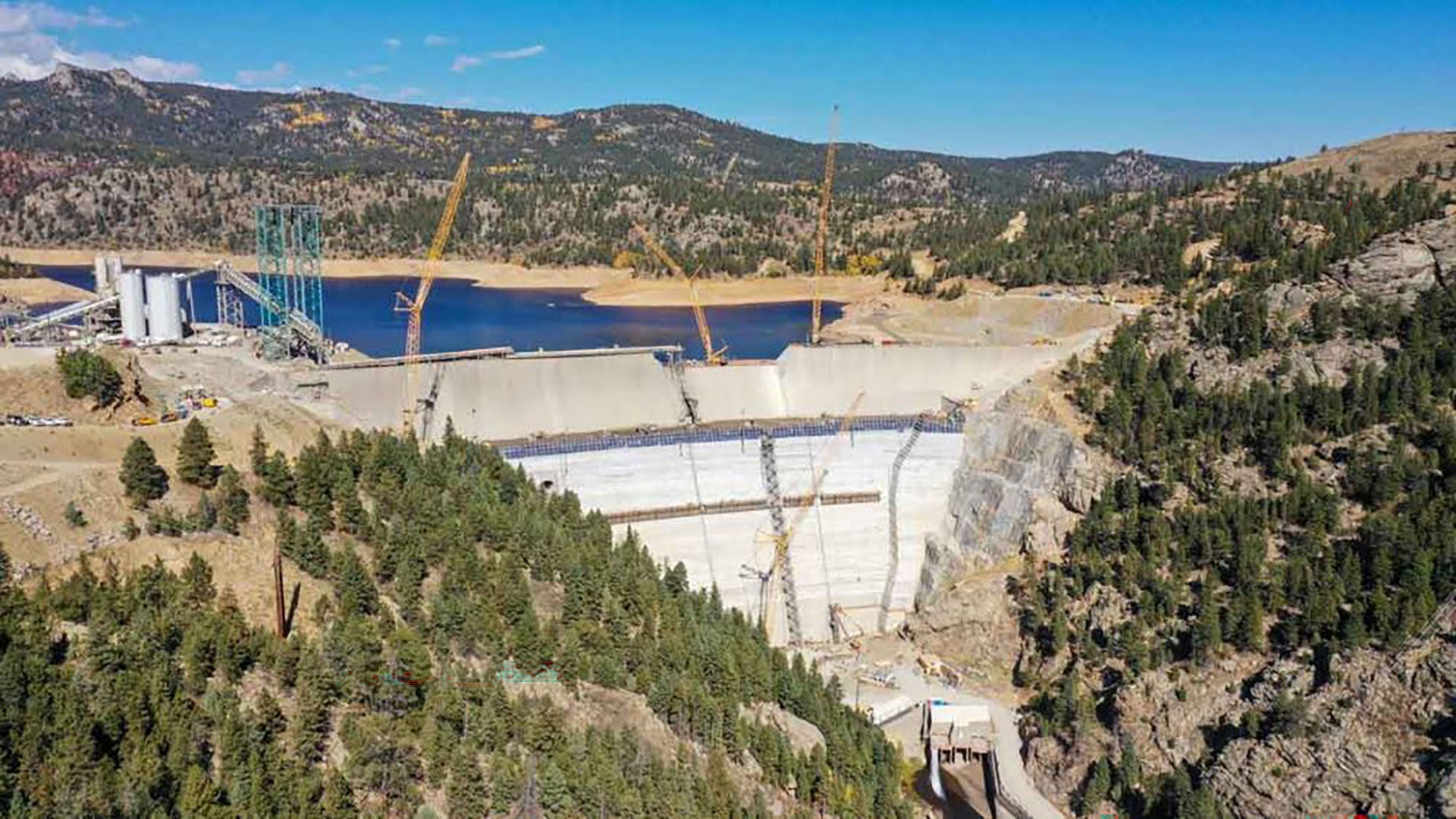 Aerial view of a dam under construction in a mountainous area, surrounded by forested hills. Cranes are visible, and a reservoir forms behind the dam.