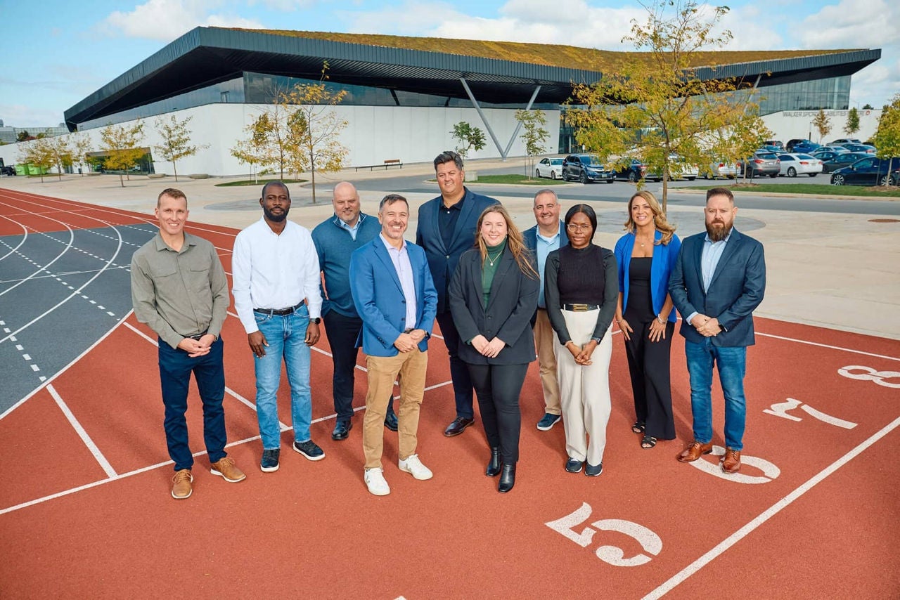 Dix professionnel(le)s debout sur une piste de course devant une installation sportive moderne, posant en toute confiance pour une photo de groupe par une journée ensoleillée.