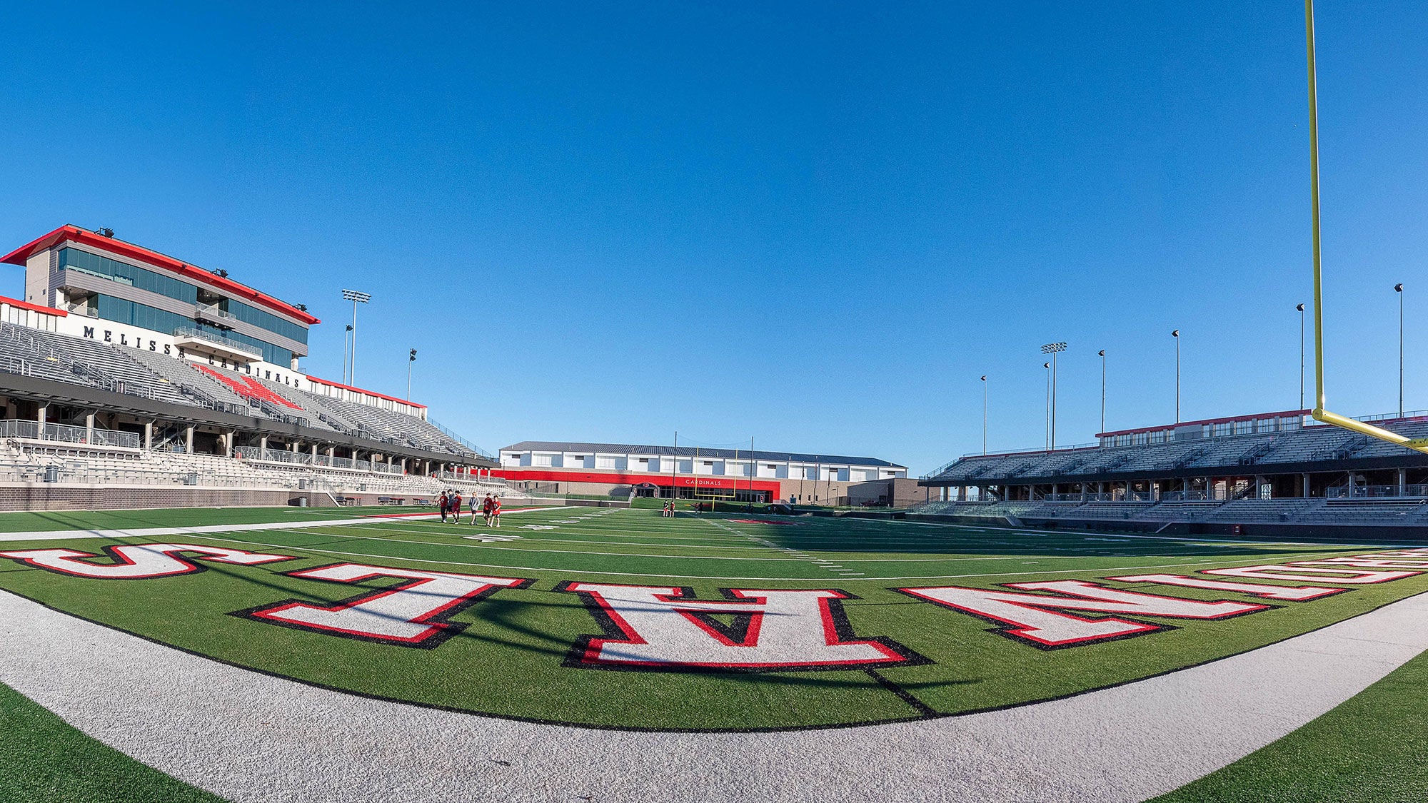 Wide-angle view of the Melissa High School football stadium and turf field under a clear blue sky, featuring "Cardinals" end zone lettering.