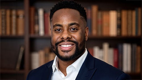 Headshot of Uchenna Nwogu wearing a navy suit jacket and open-collar white shirt in front of a bookshelf.