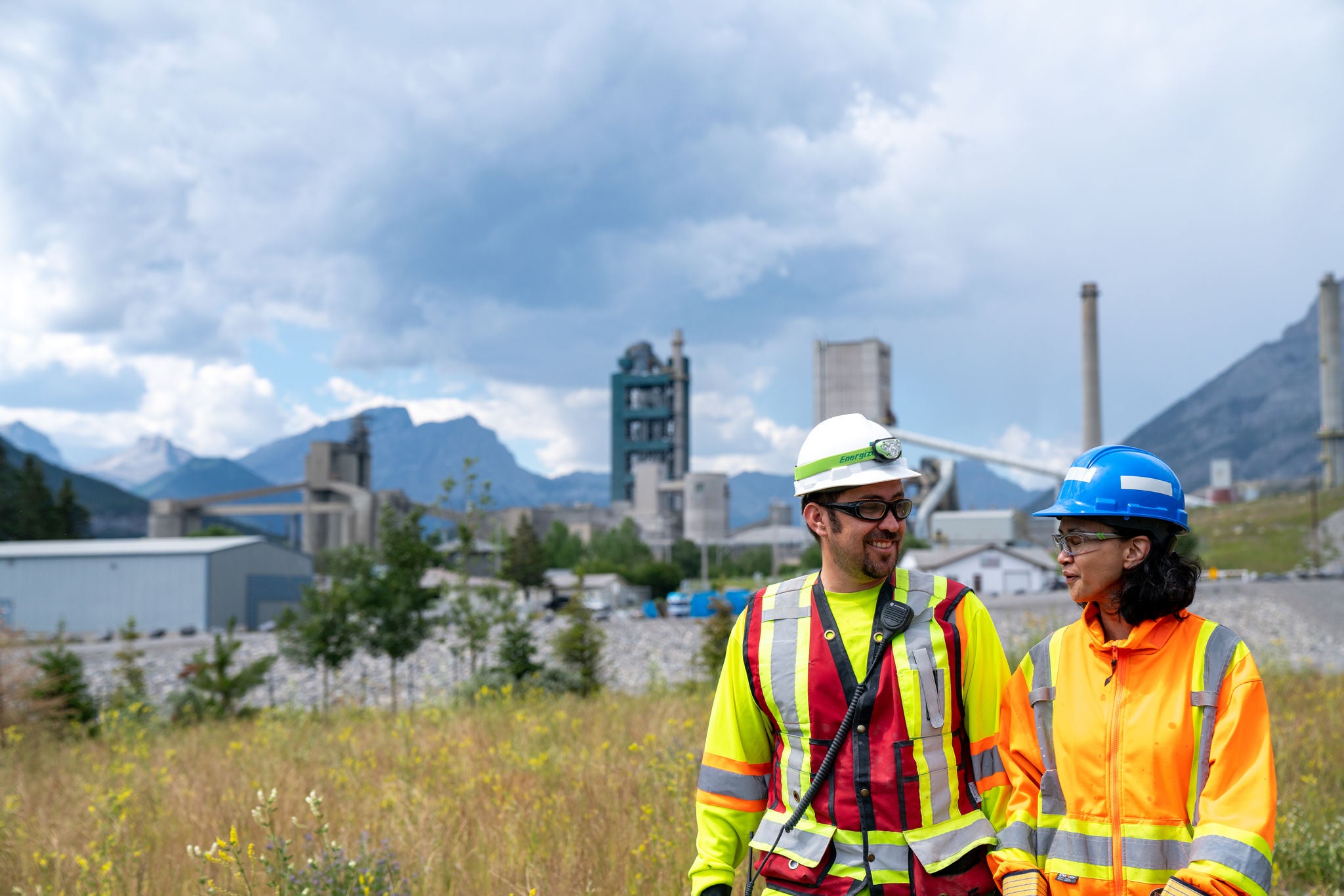 A man and a woman in personal protective gear walking in a field of flowers near a cement plant