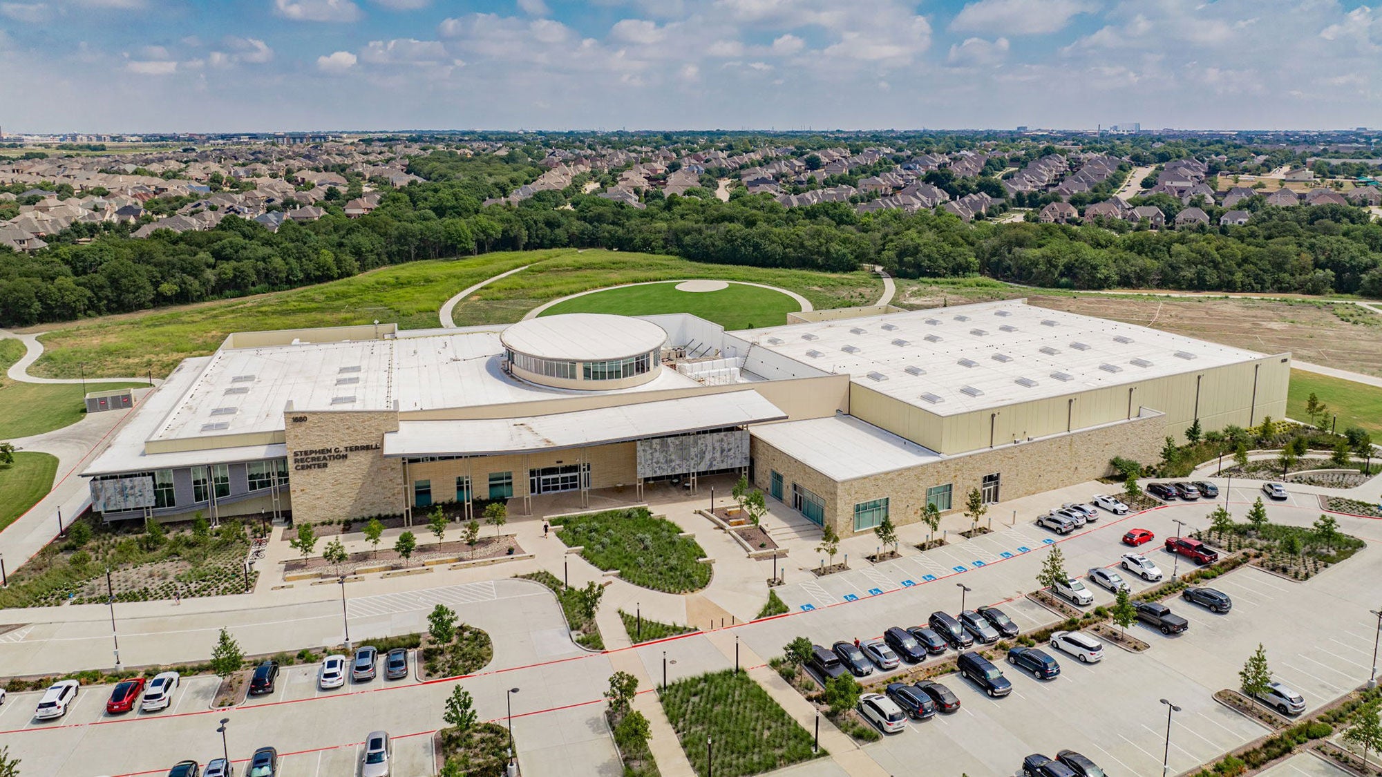High-angle aerial view of the Stephen G. Terrell Recreation Center in Allex, TX, showing the hardscape pathways by Amrize, the full building exterior and parking lot leading with surrounding green space.