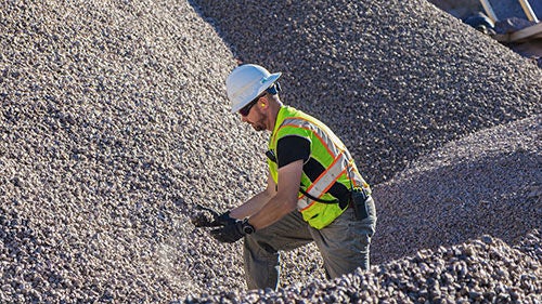 A man in protective gear standing next to a large aggregates pile