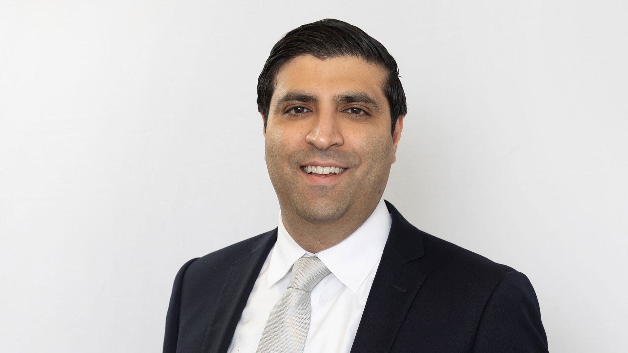 Professional headshot of Aroon Amarnani, wearing a black blazer and white collared shirt against a neutral gray background.