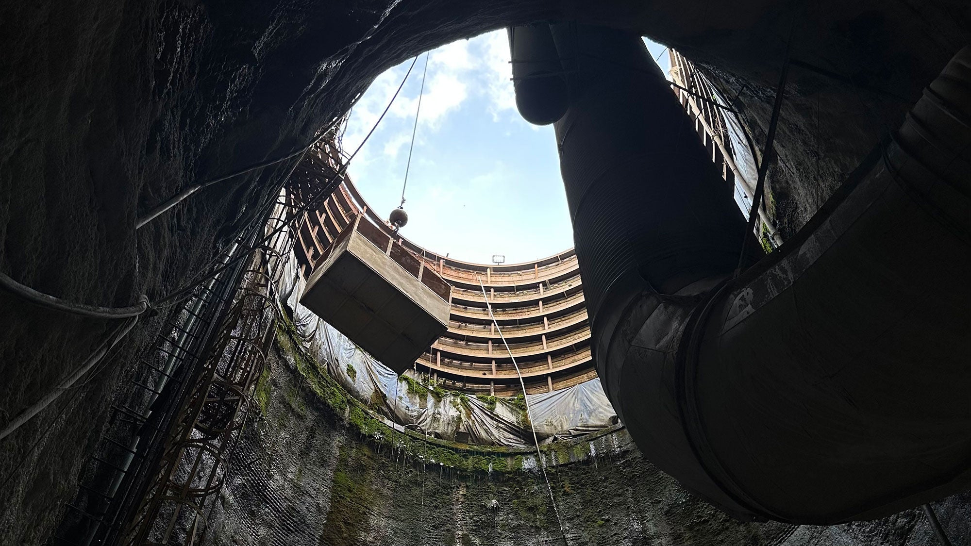 Low-angle view looking up a deep vertical construction shaft with a crane cage and ventilation pipe leading to the sky.