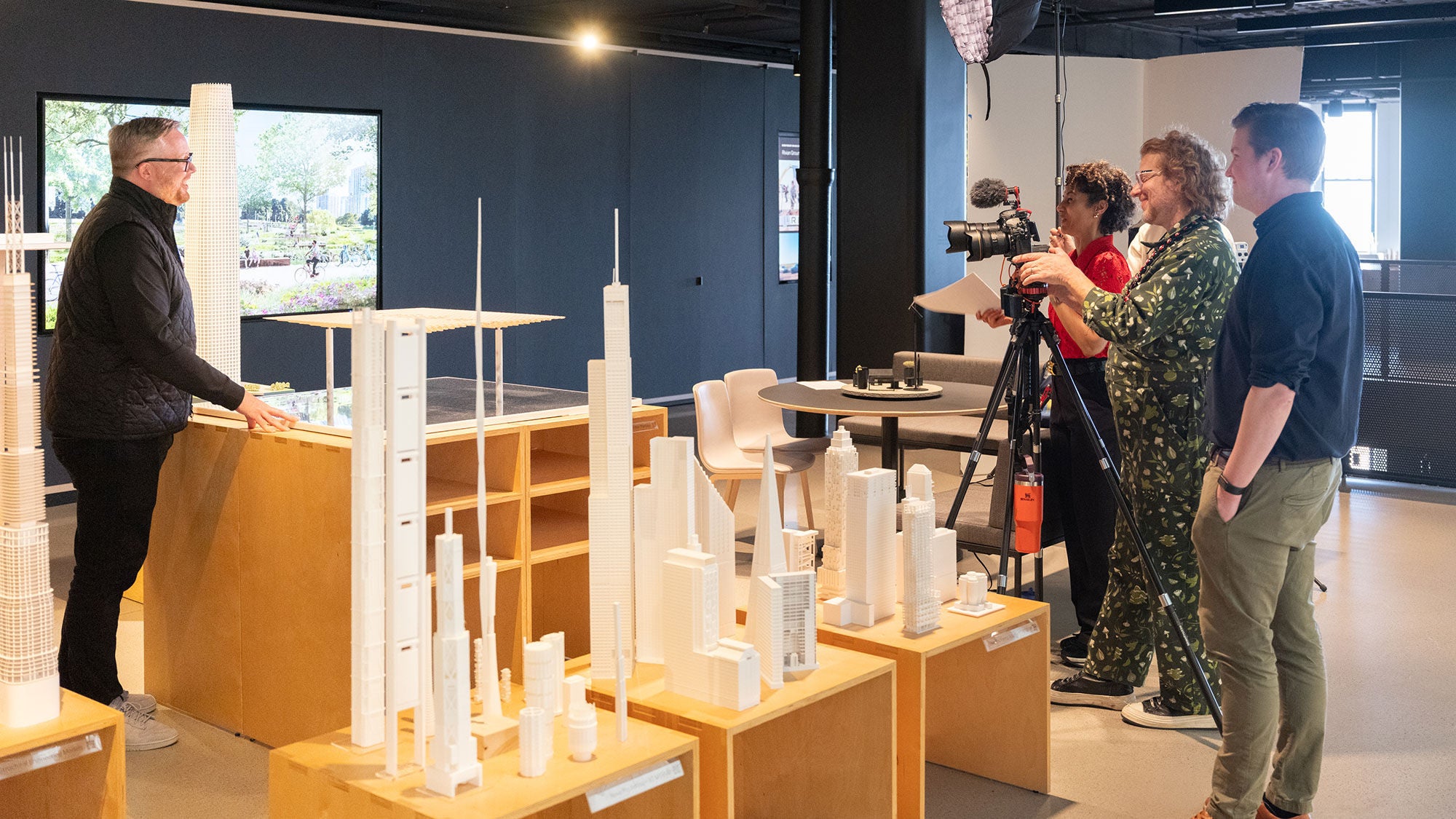 An architect is interviewed by a film crew standing behind a number of architectural models. 