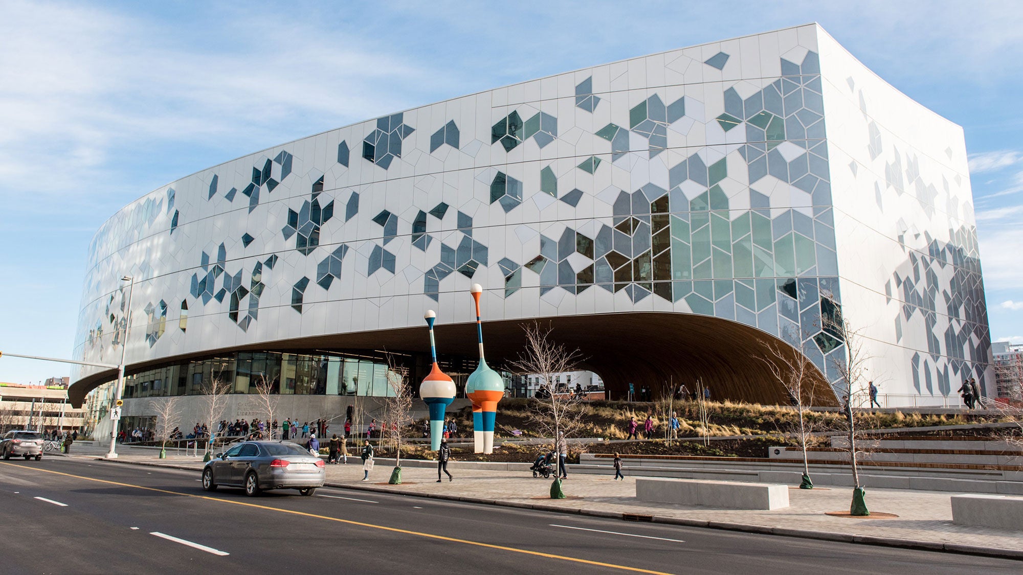  Wide exterior shot of the Calgary Central Library building featuring its geometric glass facade, large curved archway, and colorful public art sculptures in the plaza.