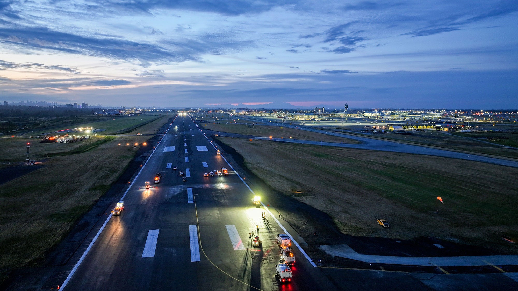 Aerial view of an airport runway at dawn, lined with maintenance vehicles and lit by floodlights. City skyline and subtle colors in the sky create a serene atmosphere.