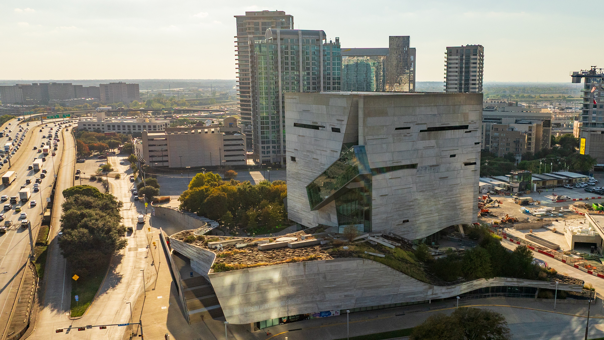 A modern, textured white building stands among downtown skyscrapers under a clear sky. Trees and urban greenery surround the area, creating a blend of nature and architecture.