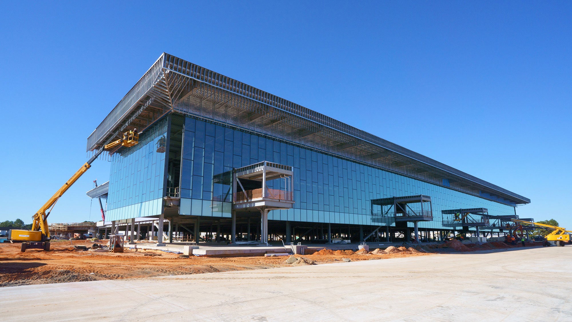 A large, modern glass building under construction on a clear day. Heavy machinery operates on red earth around the building.