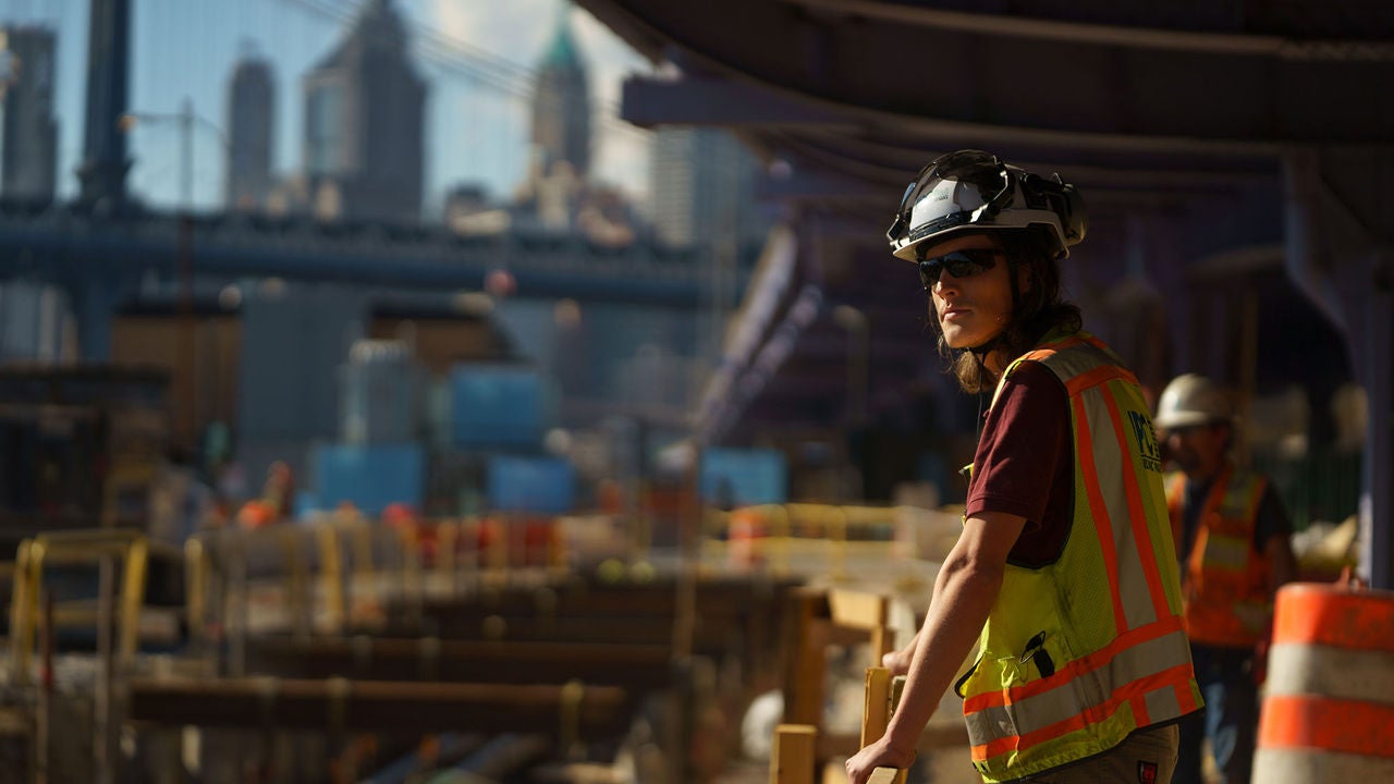 A construction worker in personal protective equipment leans against a railing at a construction site