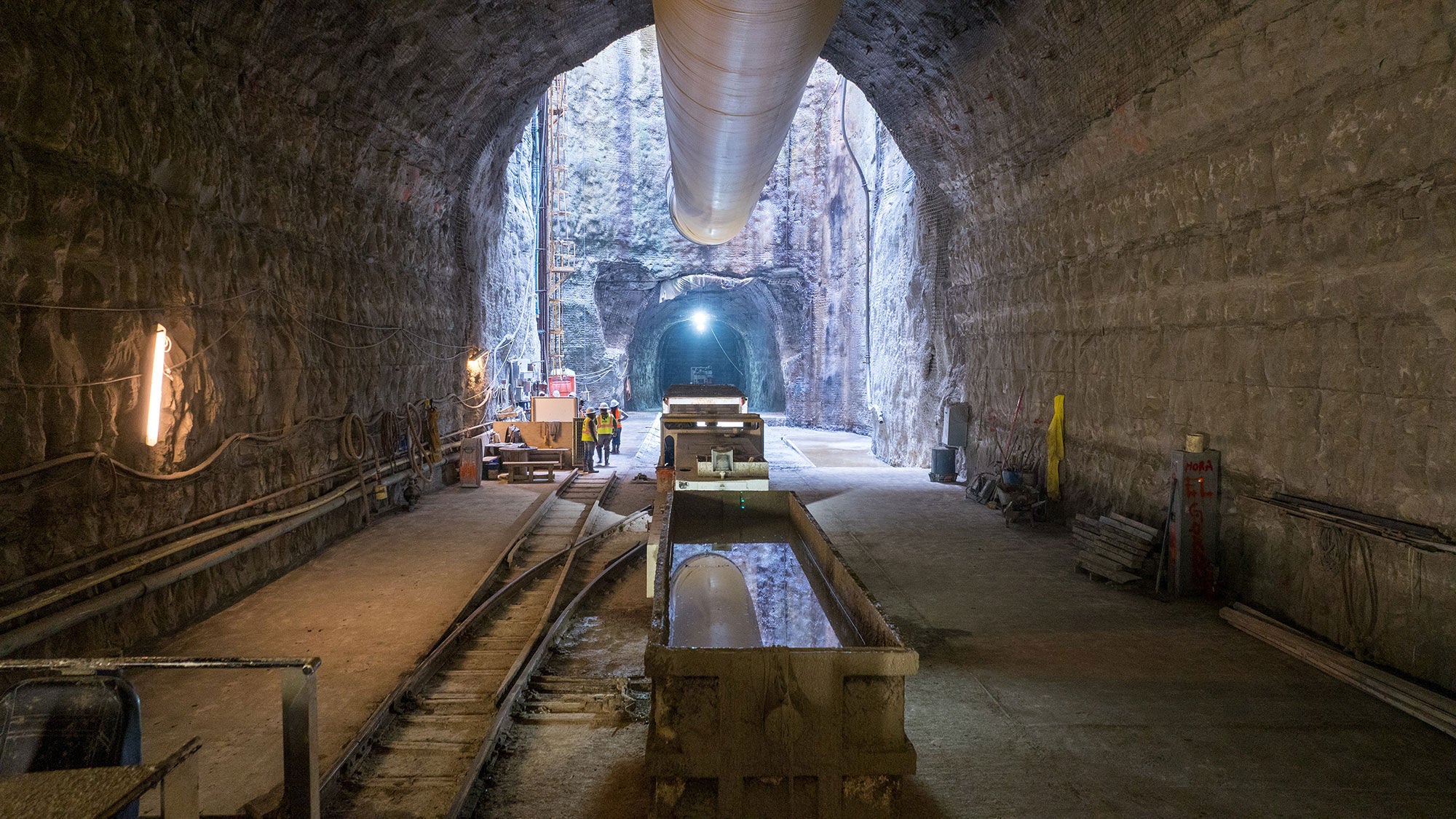 Large underground rock excavation site featuring temporary rails, ventilation tubing, and equipment inside the Mill Creek drainage tunnel project.