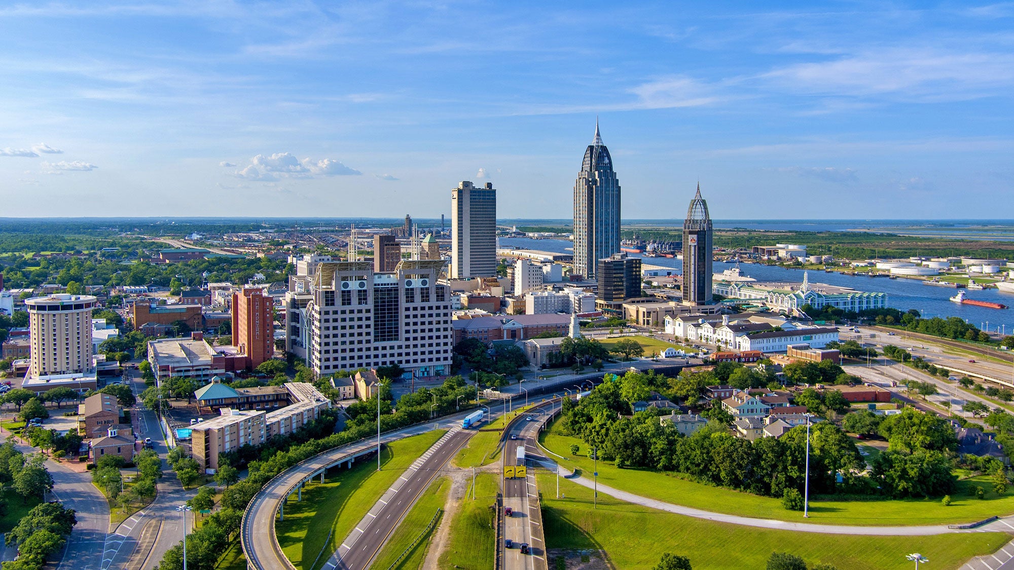 Aerial view of downtown Mobile, Alabama, featuring modern high-rise buildings, green spaces, roads and the nearby river under a blue sky.