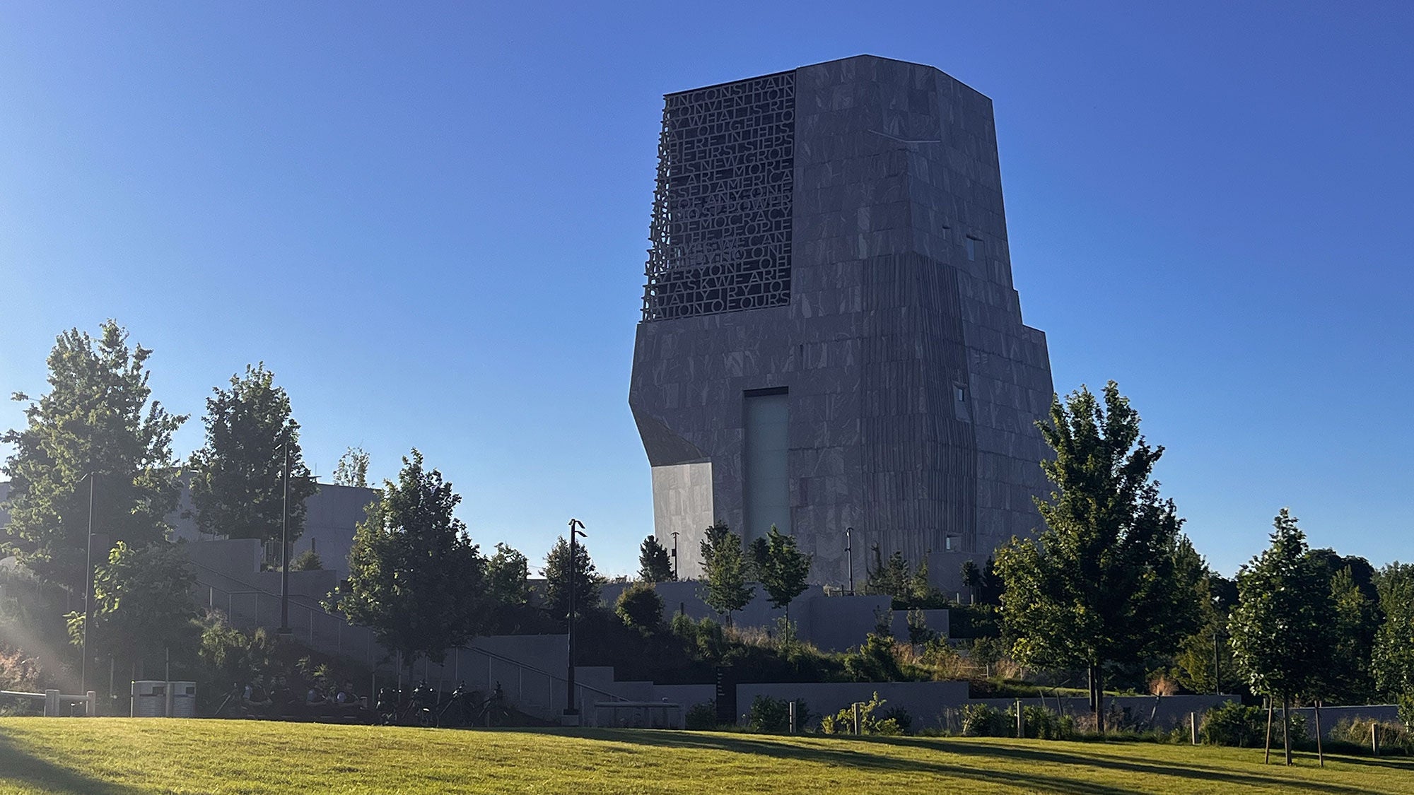 Wide exterior view of the Obama Presidential Center museum tower in Jackson Park surrounded by green lawns, trees, and landscaped campus grounds under a clear blue sky.