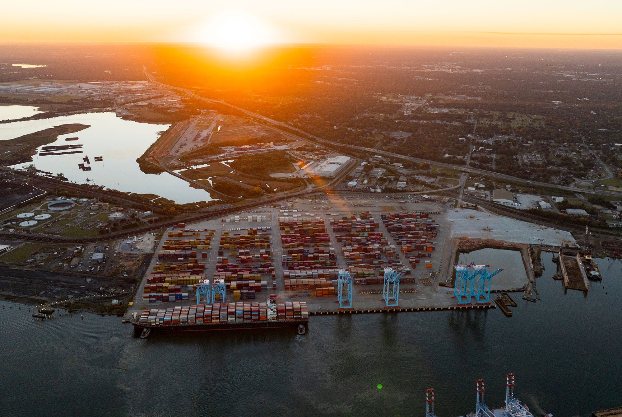 Aerial view of a busy port at sunset, with neatly arranged shipping containers and cranes along the waterfront. Warm, serene hues fill the sky.