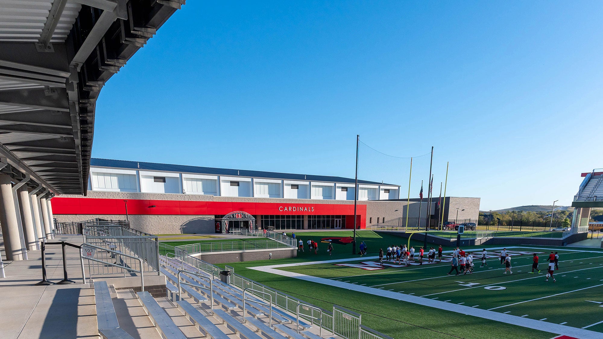A high-school athletics field shown from the stands with players on the field and a blue sky behind.