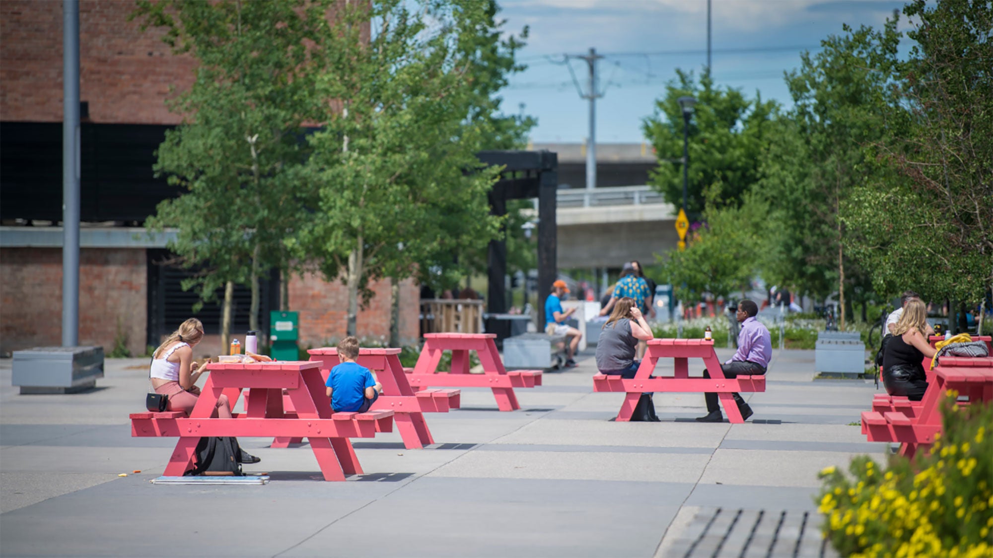 People sitting at bright red picnic tables in Calgary’s RiverWalk plaza with urban greenery.