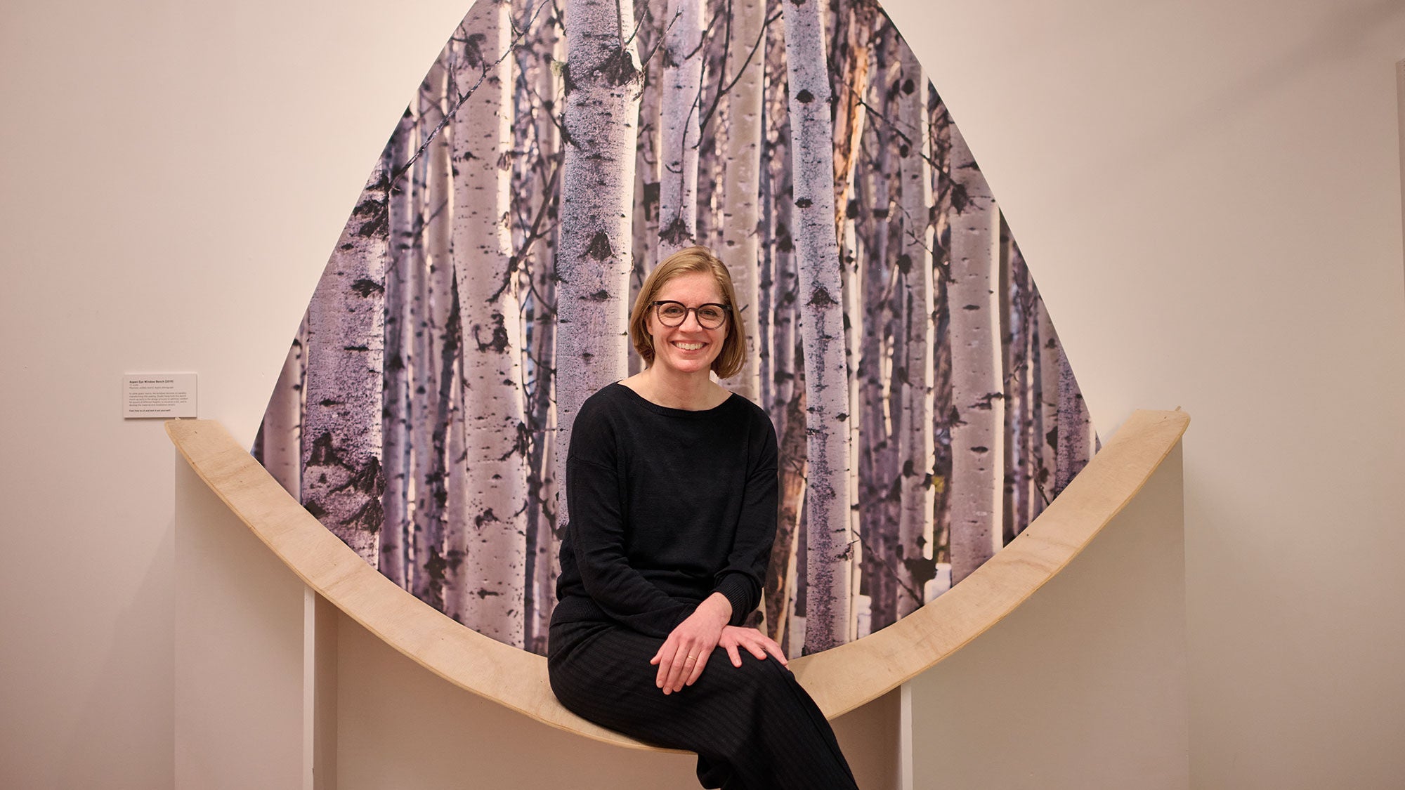 A woman sits on a curved bench with artwork featuring tree bark behind.