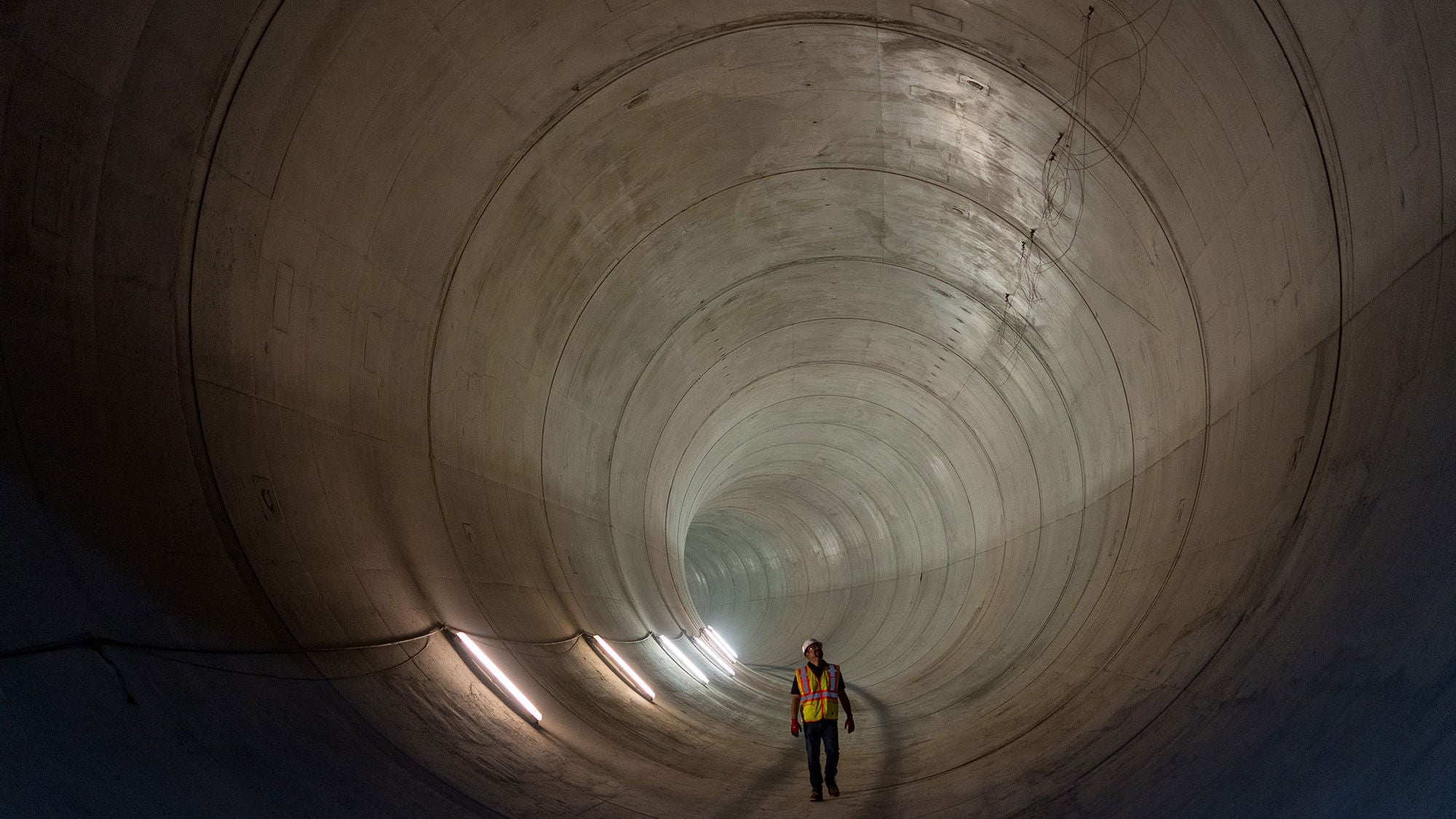 A worker walking inside the large, smooth concrete-lined interior of the Mill Creek Drainage Relief Tunnel in Dallas, Texas. 