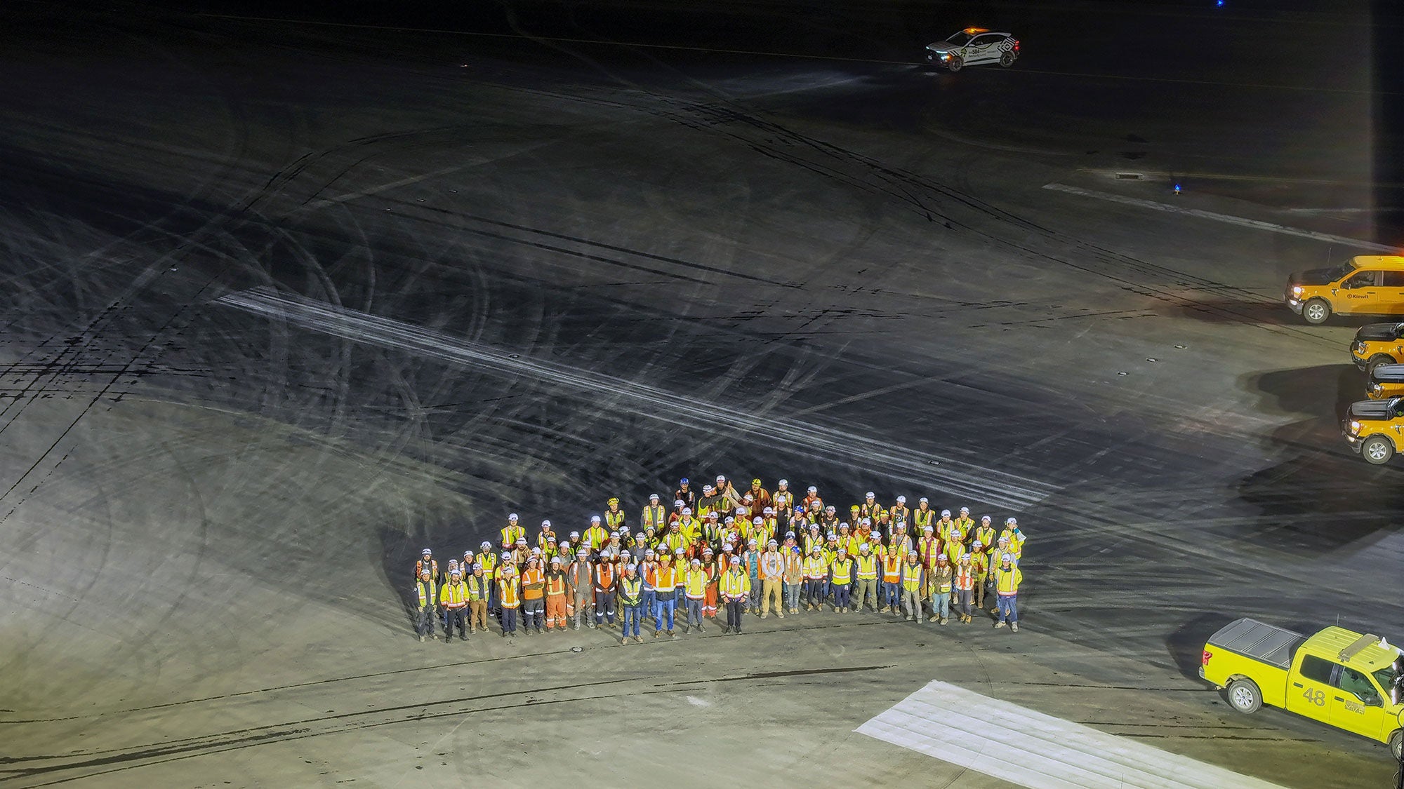 A large group of construction workers in safety gear and hard hats stand together at night on a well-lit runway. Yellow construction vehicles are parked nearby.
