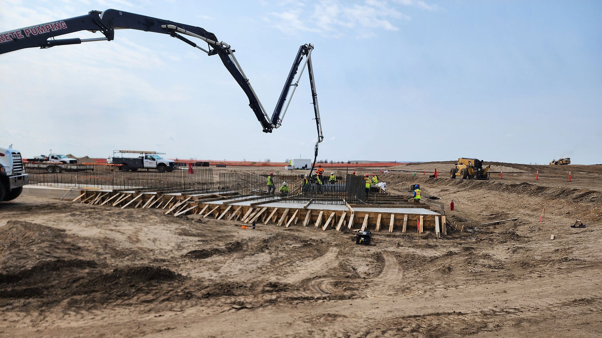  Construction workers overseeing a large concrete pour for the foundations of the Theodore Roosevelt Presidential Library, featuring a long-reach pump truck against a bright sky.