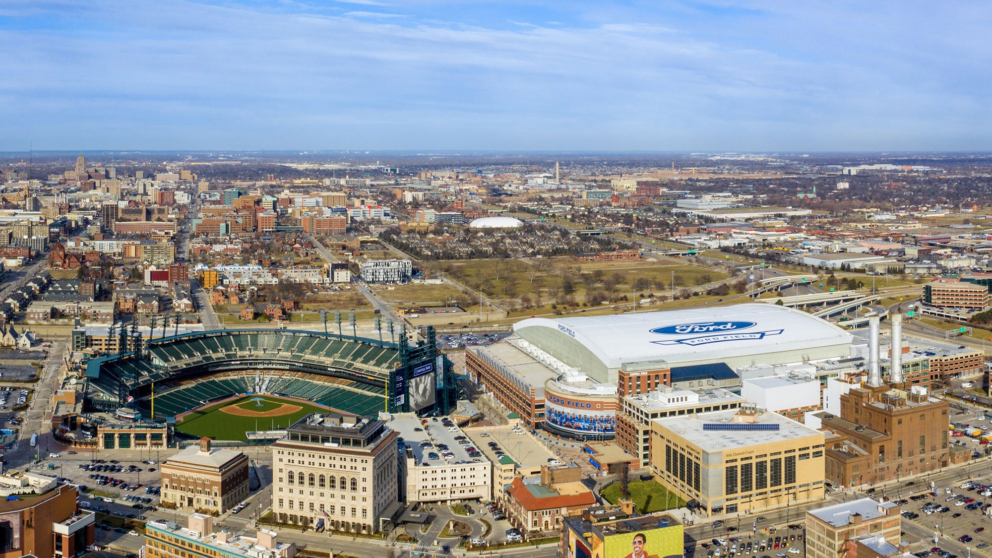 Sunny daytime aerial view of the Detroit city skyline, highlighting Ford Field stadium and Comerica Park next to downtown buildings.