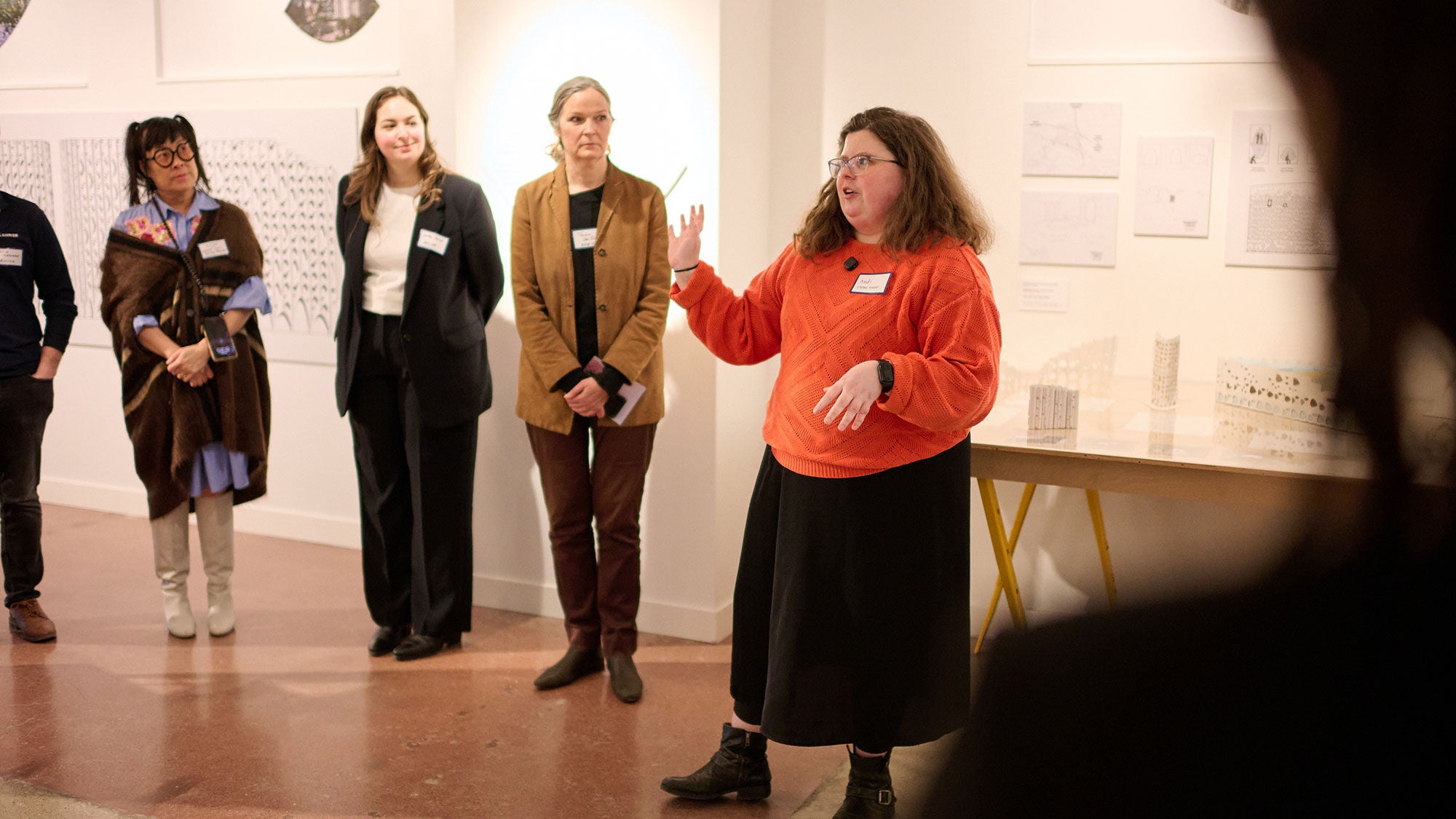 A person wearing a red sweater provides commentary to a group of listeners at an architecture studio.