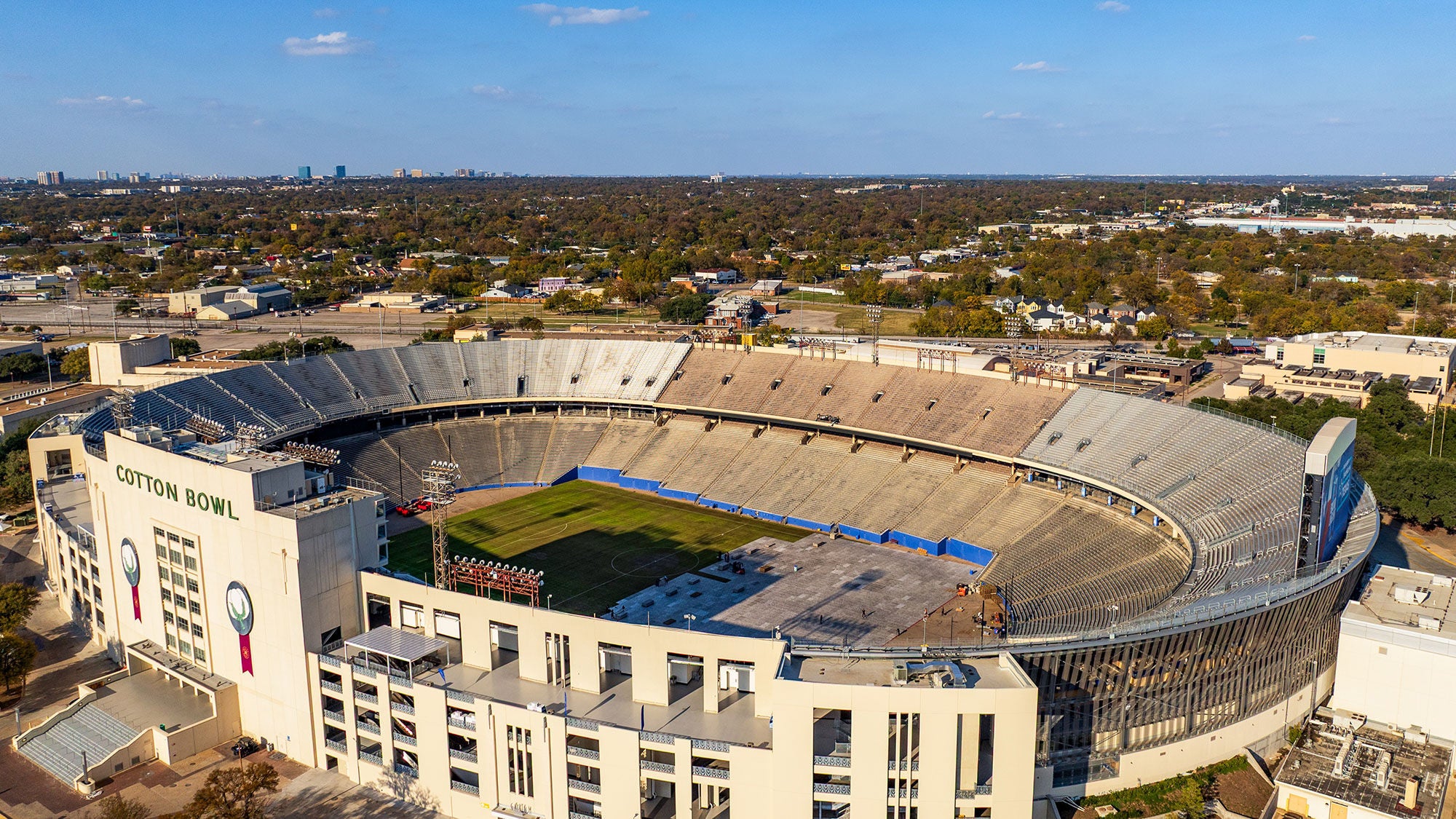 Aerial view of the historic Cotton Bowl Stadium in Dallas, Texas, showing the empty grandstands, green field and surrounding city landscape.