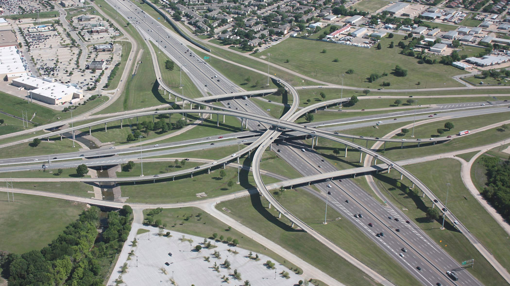 Aerial view of a large highway interchange with multiple overpasses and ramps crisscrossing in a complex pattern. Surrounding areas are residential areas and greenery.