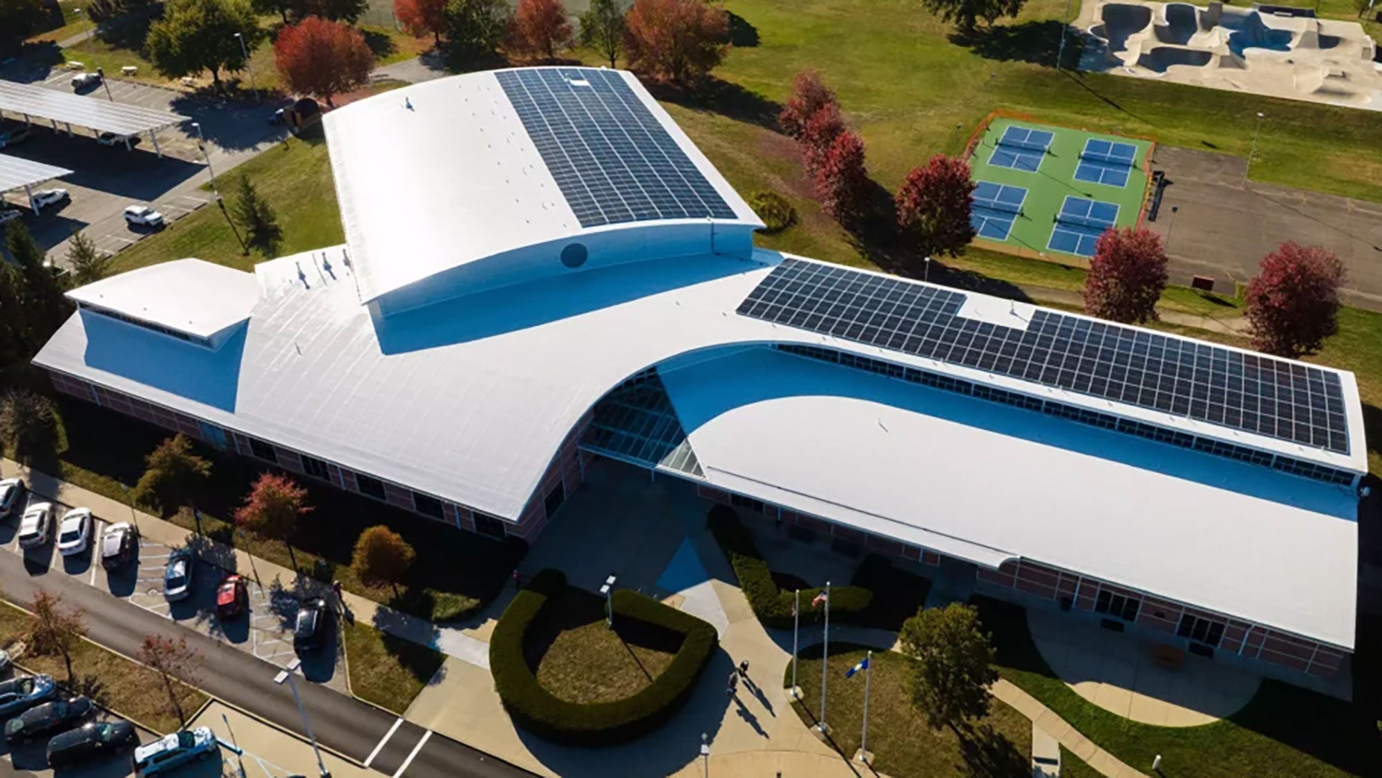 The front portion of a large, three-armed commercial building with a white roof and solar panels seen from above. Tennis courts seen behind the building.