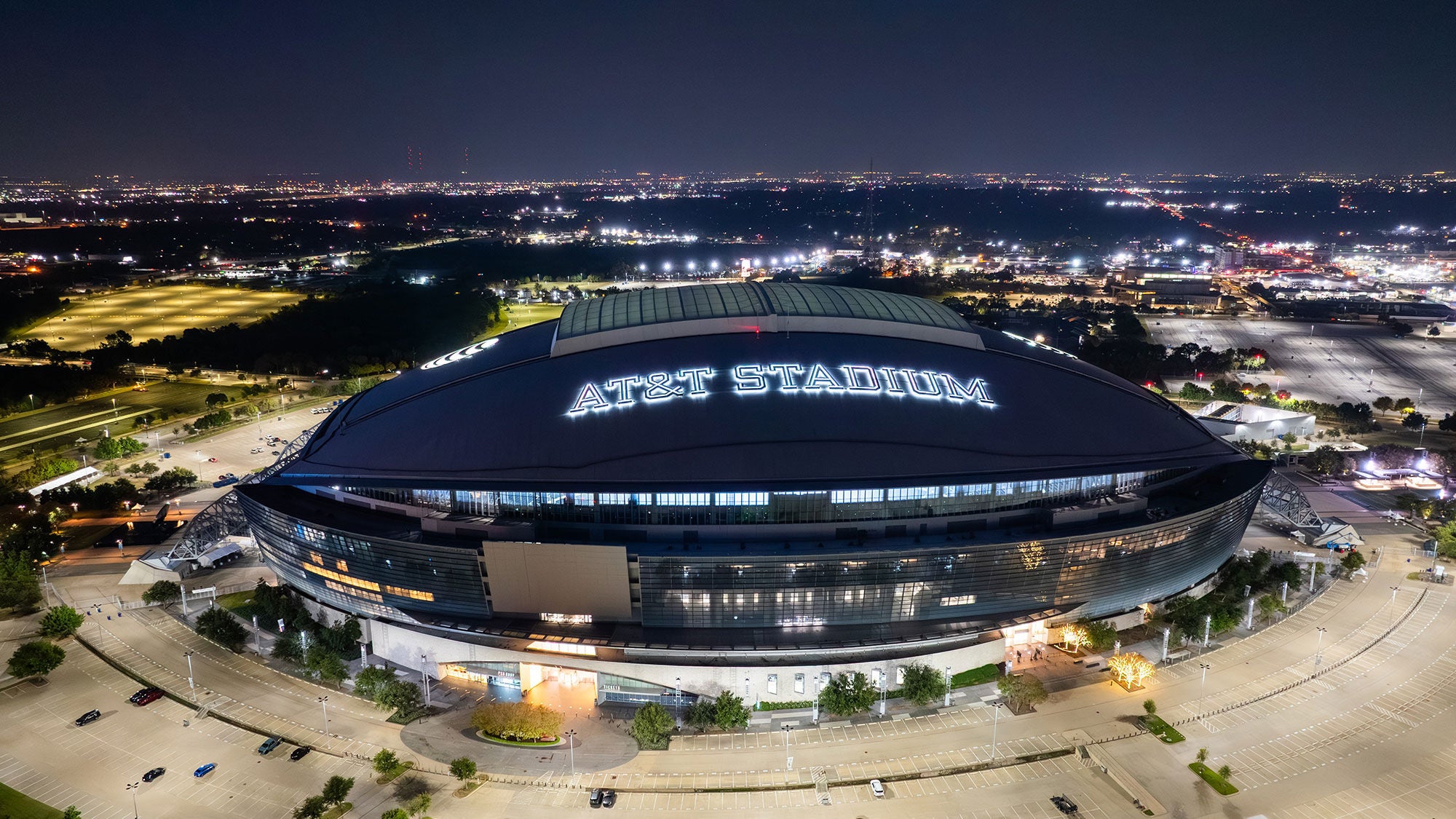 Nighttime aerial drone photograph of AT&T Stadium illuminated in Arlington, Texas, surrounded by empty parking lots and city lights.
