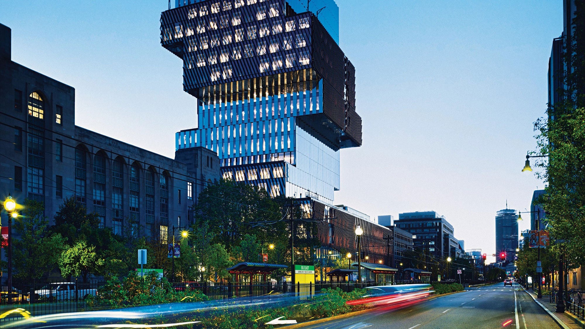 Boston University Center, with a stacked design and illuminated at dusk. Streetlights and blurred vehicle lights create a dynamic urban scene.