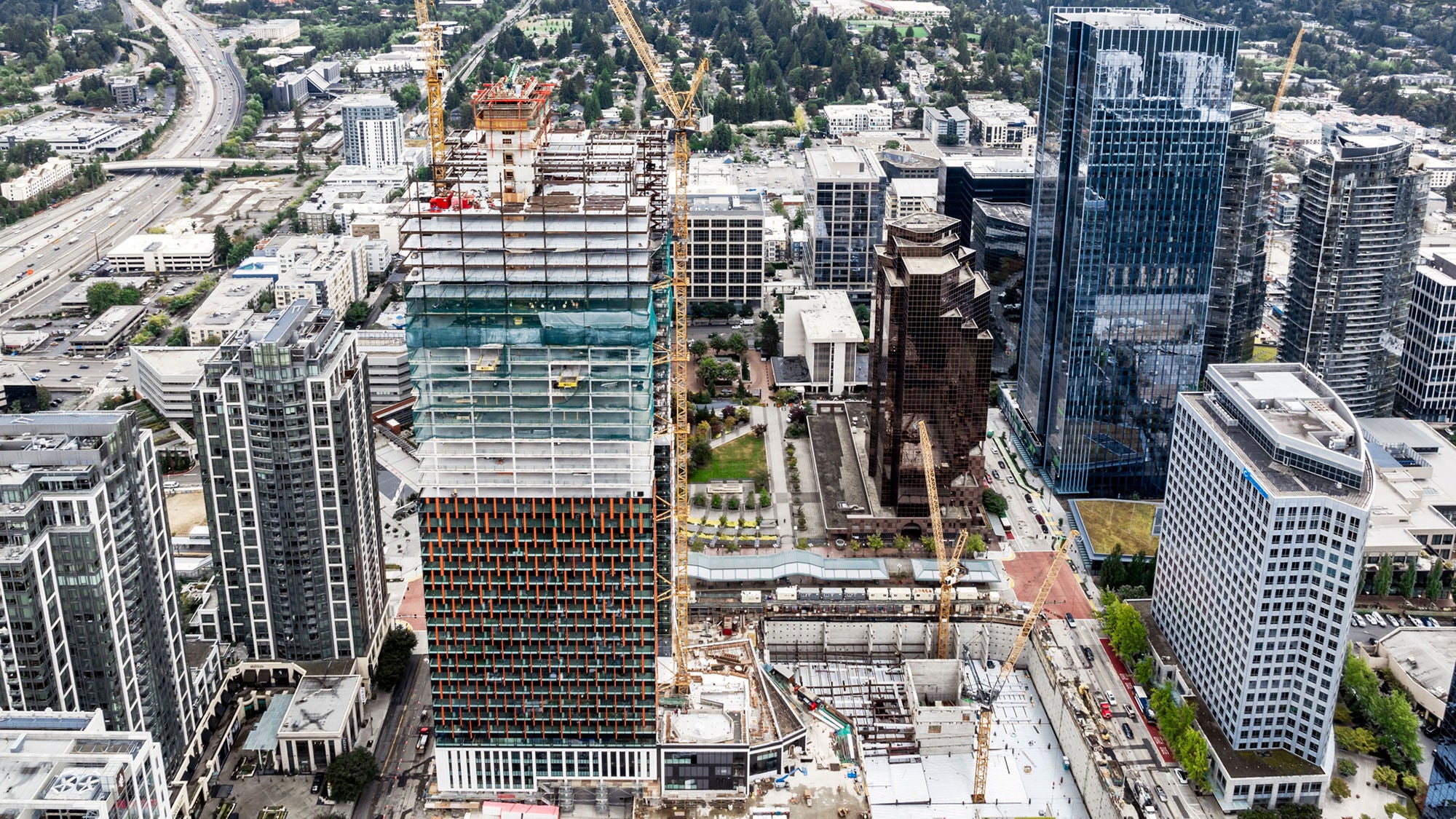 A high-rise building under construction in the middle of a city with other tall buildings around it.