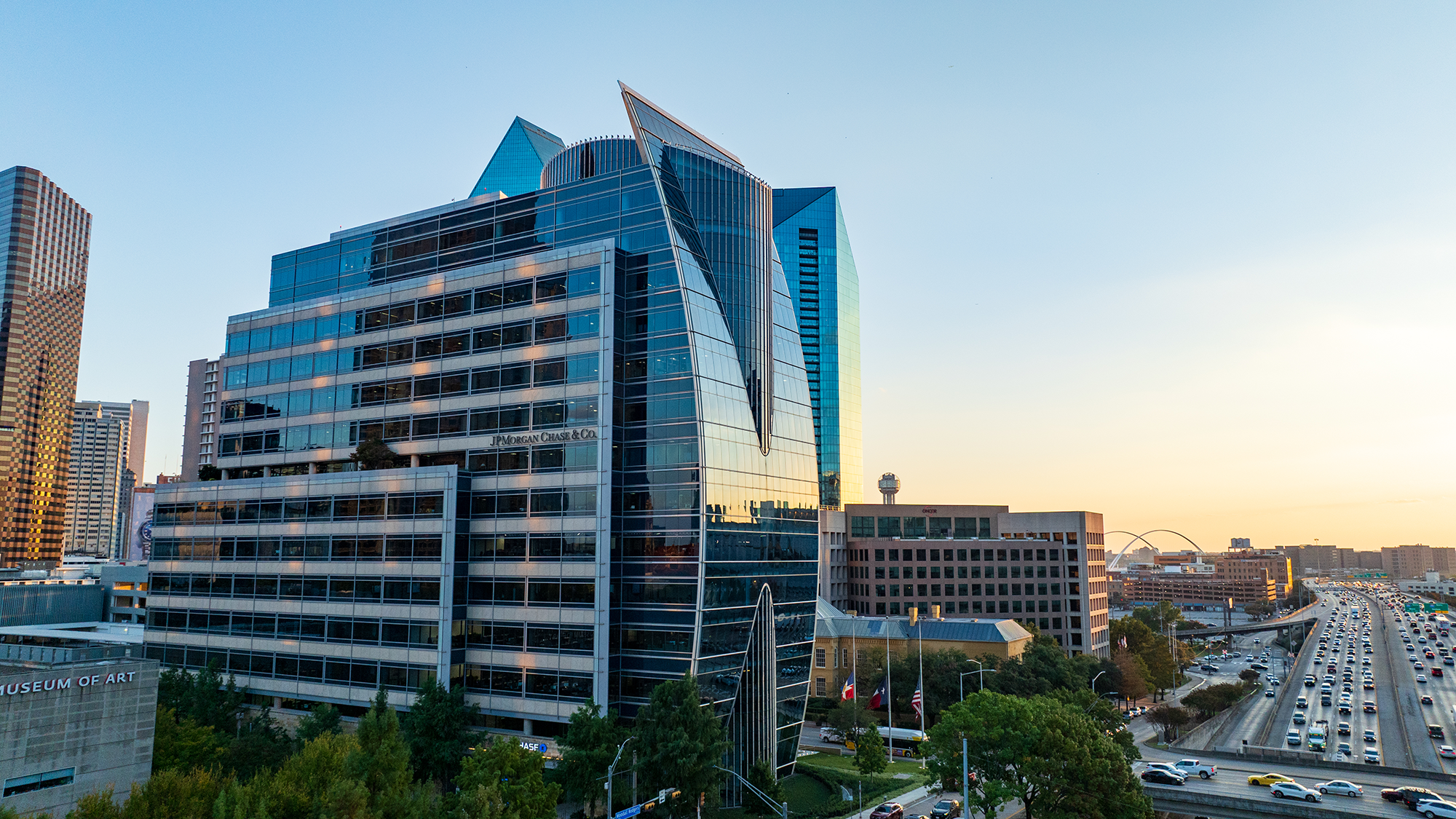 A modern glass building with a sleek, angular design reflects the sunset. Nearby, busy traffic flows on a highway, conveying an urban, vibrant atmosphere.