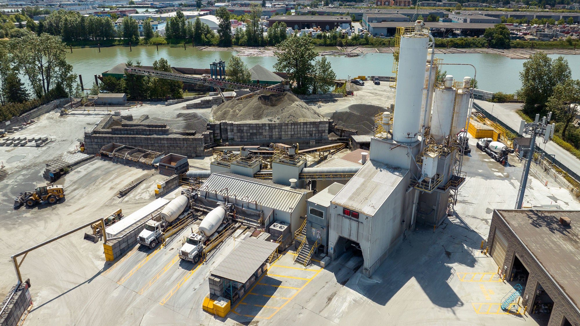 Vue aérienne d’une usine de béton au bord d’une rivière, avec des silos de stockage, de grandes piles de gravier, des camions de béton et des bâtiments industriels, avec une verdure luxuriante et des ponts en arrière-plan.