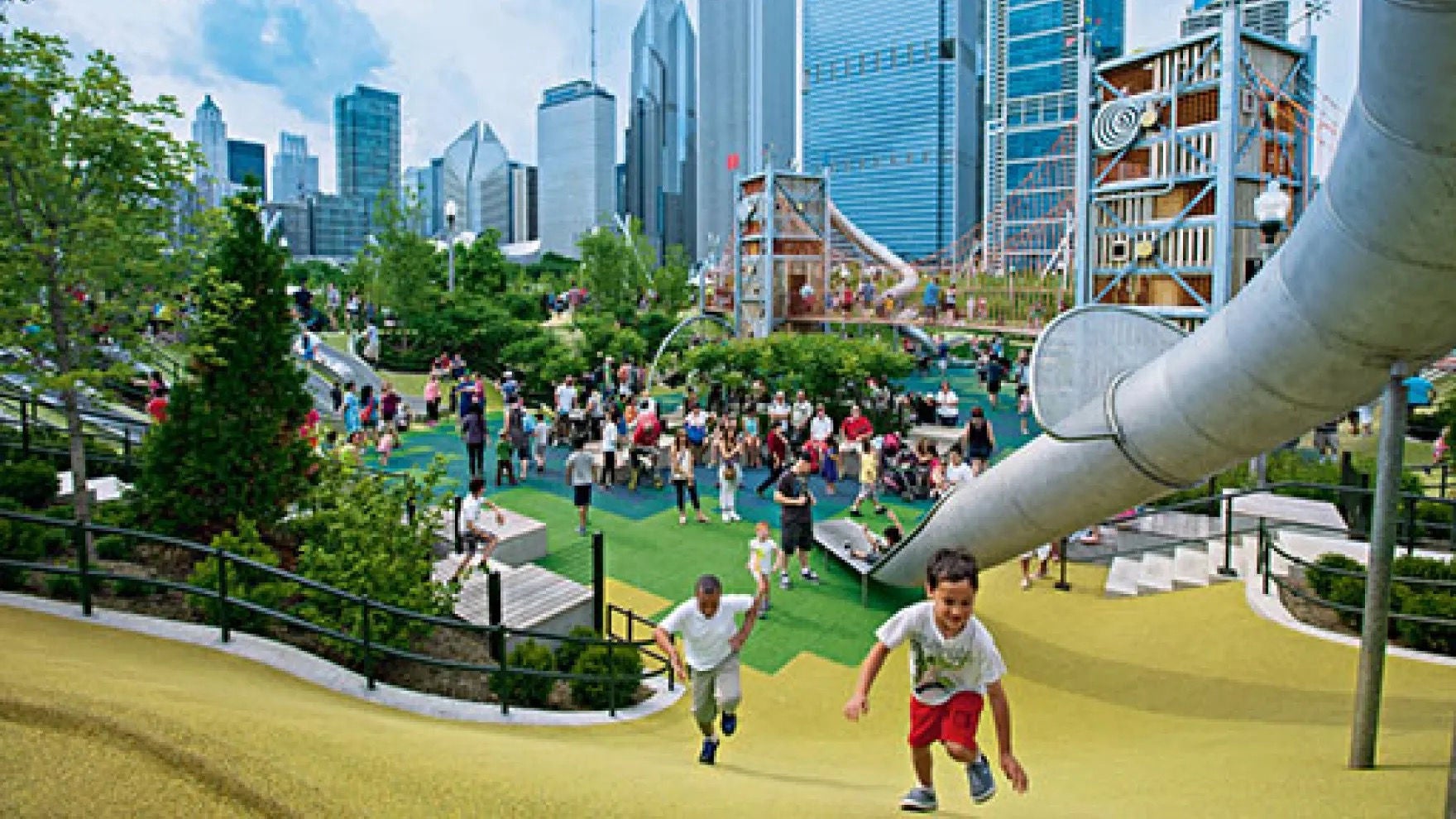 A scene of Maggie Daley Park in Chicago featuring children racing up a hill in the foreground to the top of a long slide, with many families in the background enjoying the large playground in a metropolitan setting. 