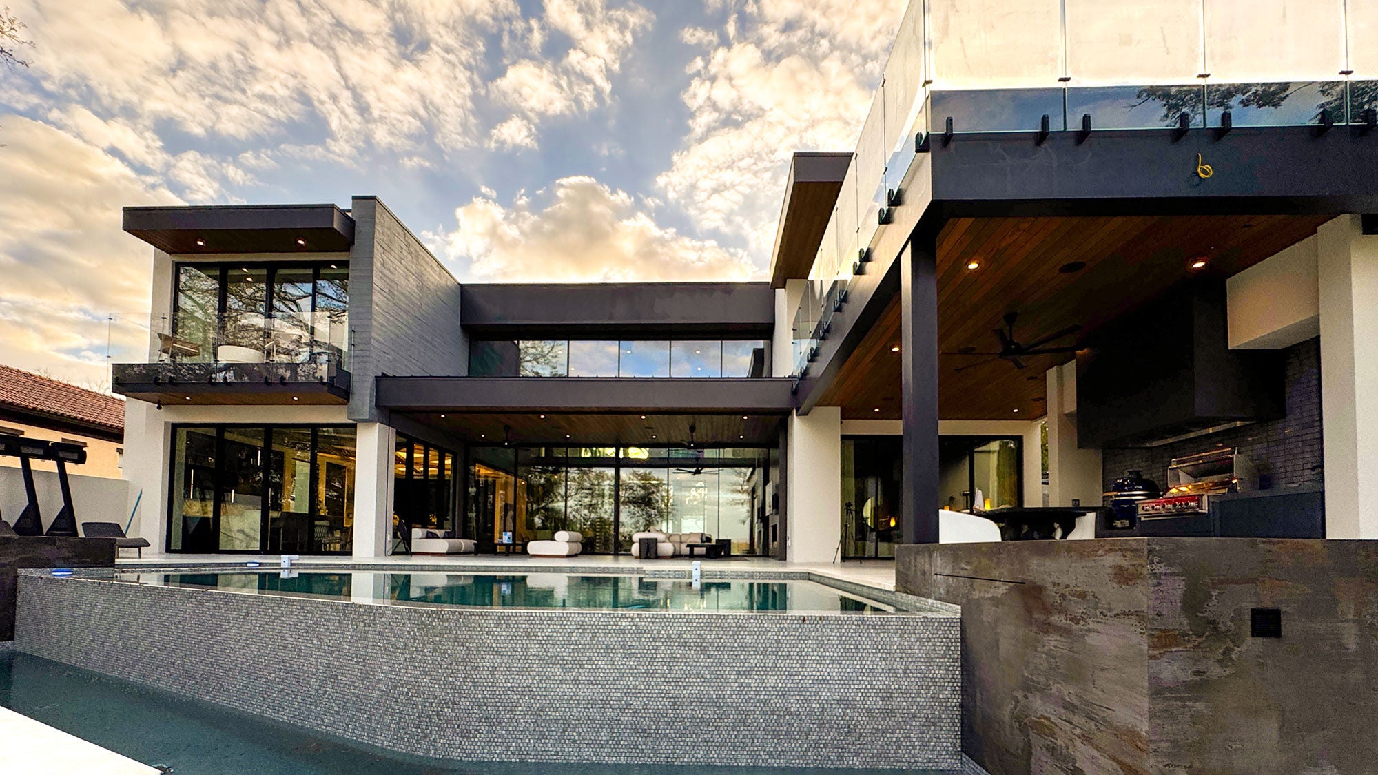 The New American Home 2026 rear facade, showing a two-storey modern residence with floor-to-ceiling glazing, a curved pool, terraced outdoor entertaining areas, fire features, and an outdoor kitchen, set among mature trees in Winter Park, Florida.