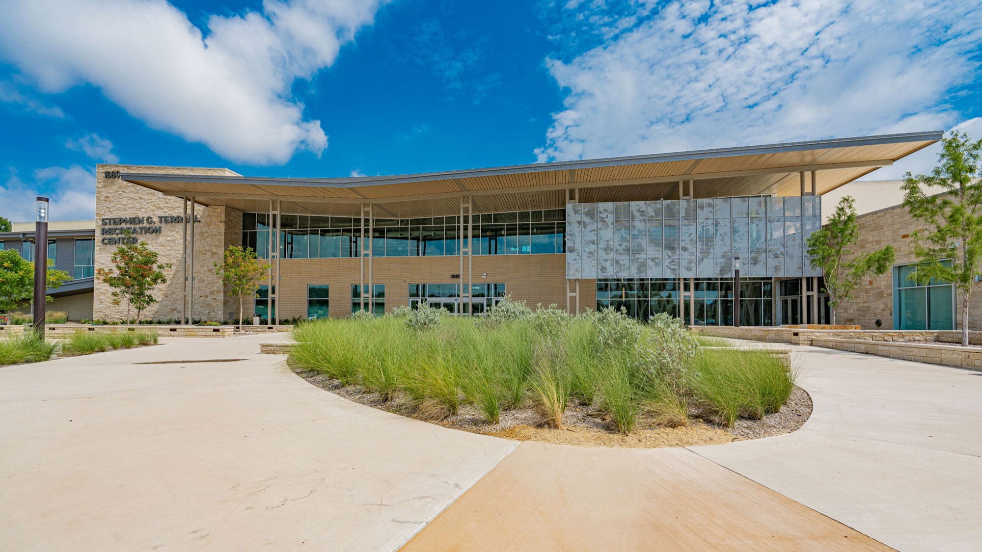 A ground–level view of the Stephen G. Terrell Recreation Center’s main entrance in Allen, Texas, featuring a curved concrete plaza and landscaped gardens with green grass.