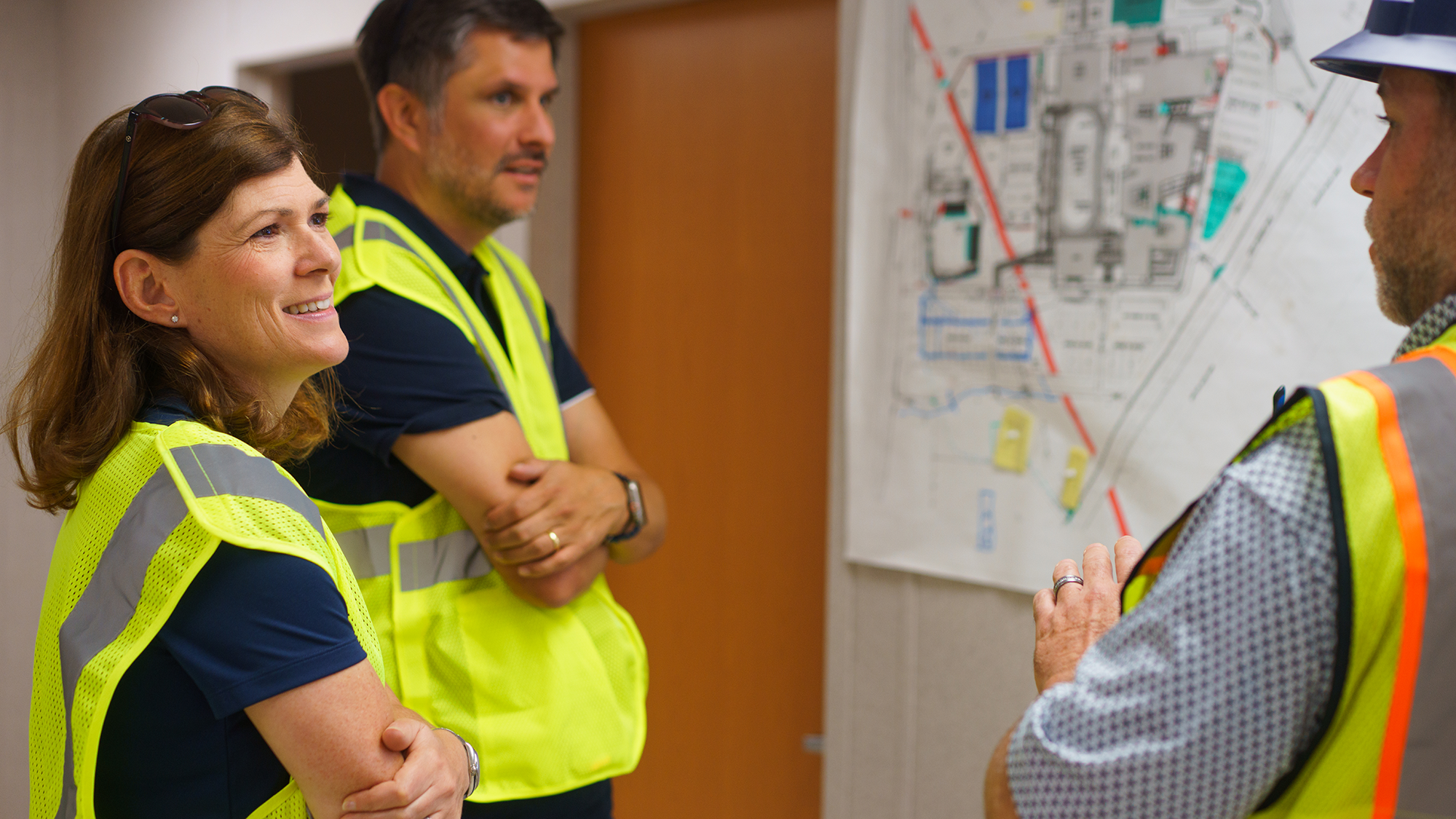 Two people in high-visibility vests stand smiling and engaged in conversation with a third person in a hard hat near a wall displaying a large site map.